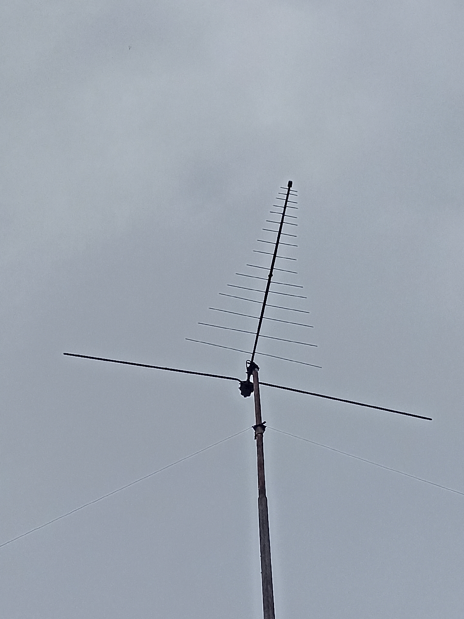 An outdoor, low-angle, full shot captures a television antenna against a gray, overcast sky. The antenna is mounted on a wooden pole that extends vertically from the bottom of the frame. The antenna itself is a Yagi-Uda design, consisting of a central boom with multiple parallel elements of decreasing length, forming a triangular shape. A horizontal element protrudes from the boom near the base. The antenna is dark, contrasting with the pale sky. There is a small dark speck, possibly a bird, visible in the upper left quadrant of the sky. The scene is devoid of any visible signs of human activity, urban environment, or geographical markers that would allow for a specific location within Mochische, Russia to be identified.