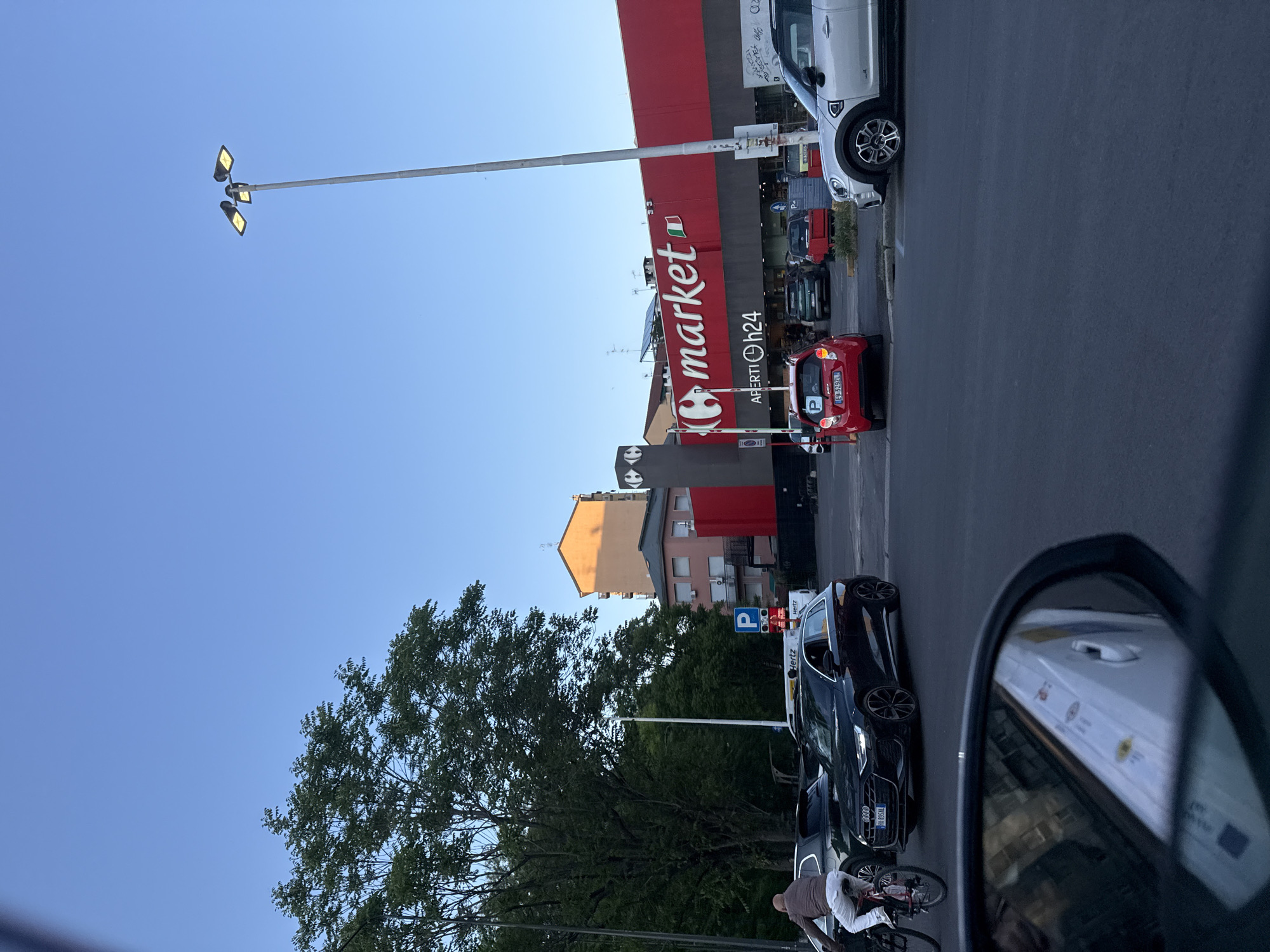 This media file captures an outdoor street scene, likely in a commercial area of Milan, Italy, as indicated by visible text and context. The photo is taken from inside a vehicle, with a portion of the side mirror visible on the right reflecting a white van.
The central and most prominent feature is a supermarket with a distinctive red facade, prominently displaying the word "market" in white letters, accompanied by a small Italian flag logo. Below this, the text "APERTI h24" (Open 24h) is visible, indicating its operating hours. A tall, modern street light pole with three upward-facing lamps extends from the foreground towards the supermarket building and into the clear blue sky.
The scene includes a dark asphalt road and parking area. Several vehicles are parked: a silver Mini Cooper, a black Audi with the license plate "HA 895AL", and a small red microcar displaying the license plate "FB 3292L". The "Hertz" logo is visible on the Audi and a nearby sign, suggesting car rental services in the vicinity. A blue parking sign with a white "P" is also present.
On the left side of the image, a large, leafy green tree dominates the midground, partially obscuring other buildings in the background which feature varied colored facades, including yellow and pink. In the bottom left, a person on a bicycle is partially visible next to the parked Audi.
The weather appears clear and sunny, with a bright blue sky, suggesting it is daytime. The overall impression is that of a typical urban or suburban commercial zone with regular daily activity. Besides the main texts, faint and less legible texts like "CRED", "MOSH", "Π", "33", and "D" are also discernible through OCR.
