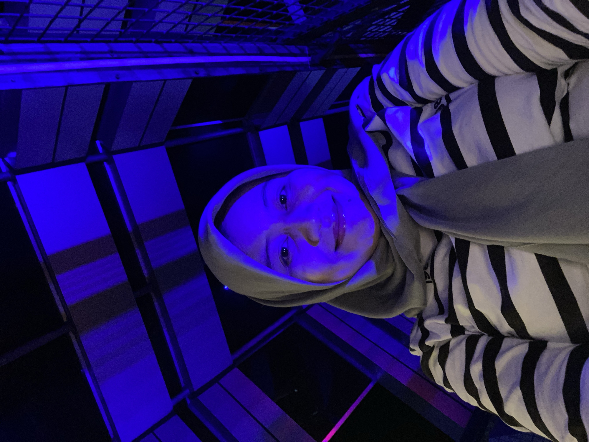 A young woman wearing a gray hijab and a black and white striped shirt is taking a selfie. She is illuminated by blue and purple lights, suggesting she is in an area with artistic or decorative lighting. The background appears to be an outdoor structure with metal beams and netting, possibly a part of an attraction or building in Banjarbaru, Indonesia. The overall atmosphere is moody and vibrant due to the colored lighting.