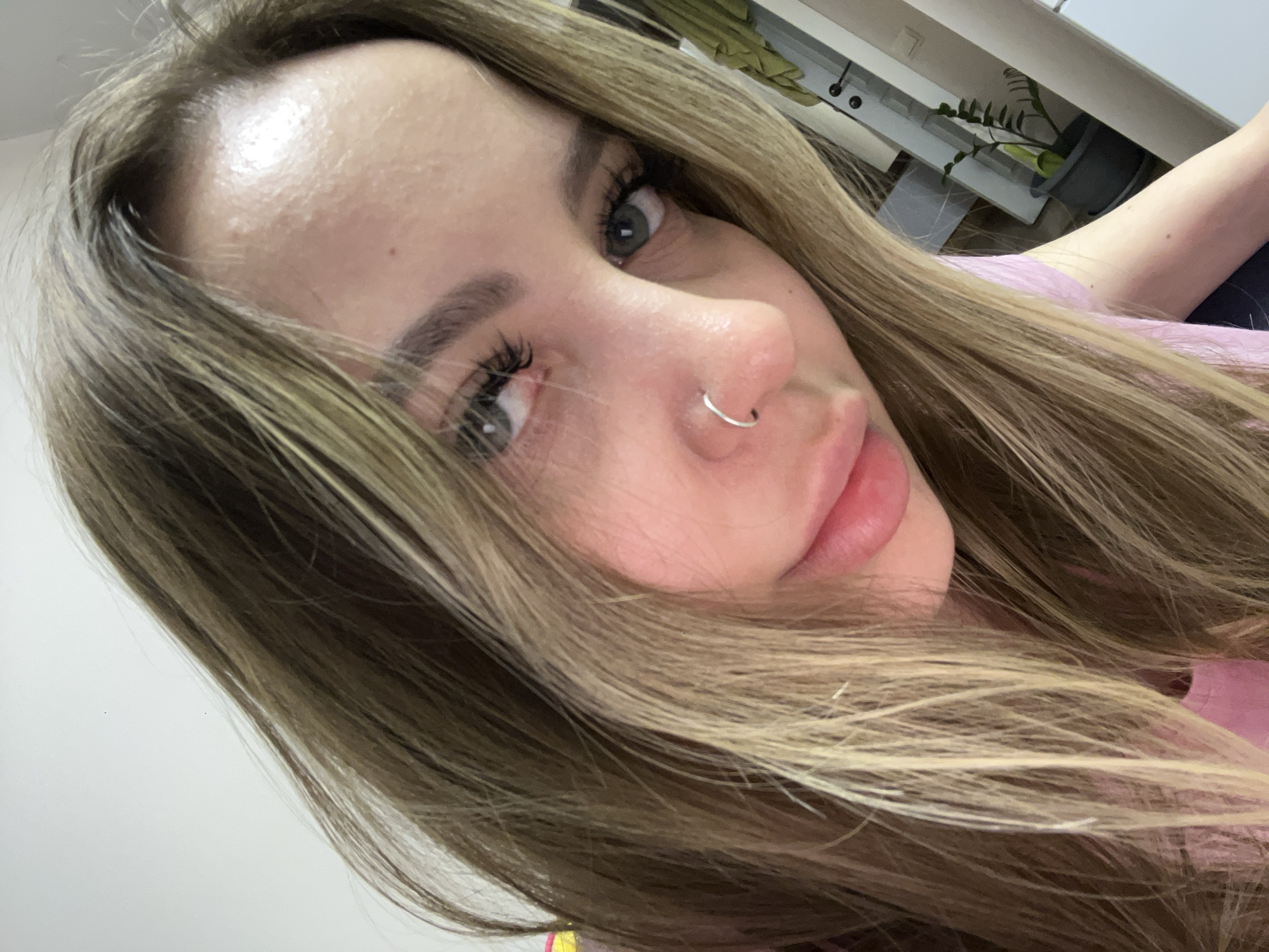 A close-up, eye-level selfie shows a woman with light brown and blonde highlighted hair, cascading across her face and shoulders. Her gaze is directed towards the camera with green or hazel eyes, accentuated by long, dark eyelashes. She has defined eyebrows and subtle pink lip makeup. A silver hoop nose ring is visible on her right nostril. Her forehead shows some texture from the close-up perspective.
The woman is wearing a light pink garment, partially visible on her shoulder and arm. In the background, suggesting an indoor setting, elements of a room are discernable. These include what appears to be a light-colored desk or shelf, and a dark plant pot with green foliage. The scene implies a casual environment, possibly a home or office in Krasnodar, Russia. The lighting is bright and even, indicating an indoor shot without specific time-of-day cues. No visible text is present in the image.