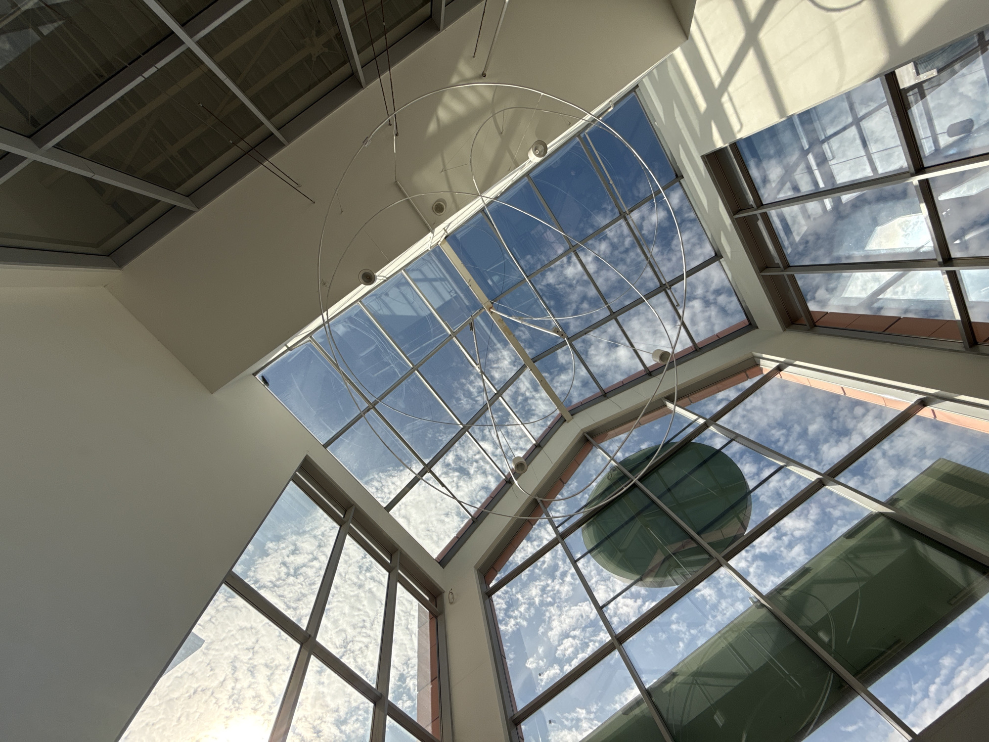 The image depicts a view upwards into a modern atrium or interior space with a large, multi-paned glass ceiling. Sunlight streams through the glass, illuminating a sky filled with white, fluffy clouds. A large, green, spherical object is visible in the reflection and through the glass, possibly part of the building's architecture or a decorative element. A contemporary, circular light fixture hangs from the ceiling. The interior walls appear to be painted white or a light neutral color, with some sections showing orange-toned accents. The overall impression is one of spaciousness and natural light. This scene is located within the architectural complex of the Bielany Wrocławskie shopping center, specifically within its interior atrium. No people are visible in the image.