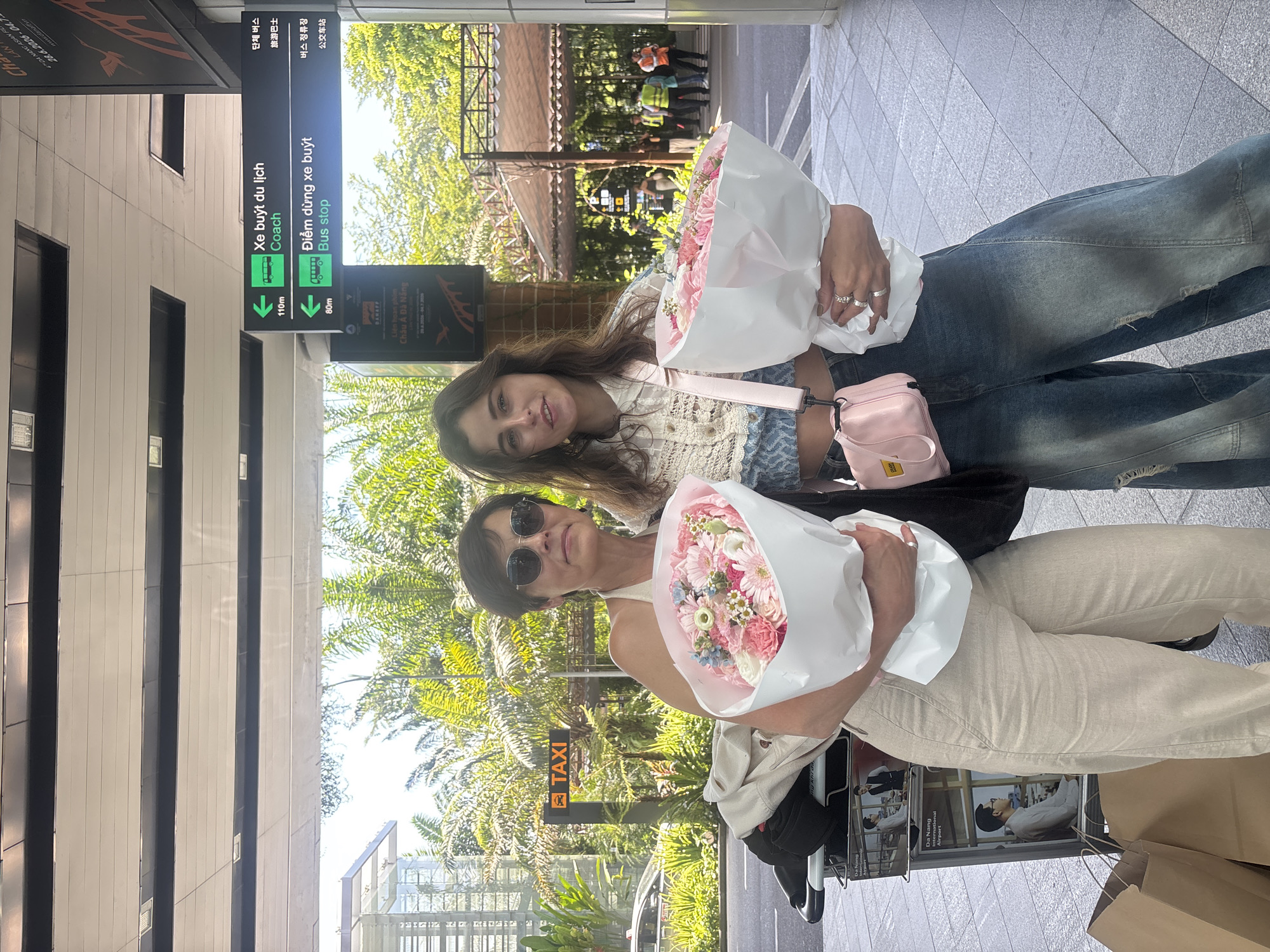 Two women are standing next to each other, holding bouquets of pink and white flowers. The woman on the left has short brown hair and is wearing sunglasses and a beige outfit. The woman on the right has long brown hair and is wearing a white and blue patterned top with denim jeans. Both women are smiling. In the background, there is signage indicating directions to a bus stop and taxi stand, suggesting the location is an airport or transportation hub in Đà Nẵng, Vietnam. The environment appears to be outdoors, with trees and a building visible. There are also luggage carts and bags present, reinforcing the travel context.