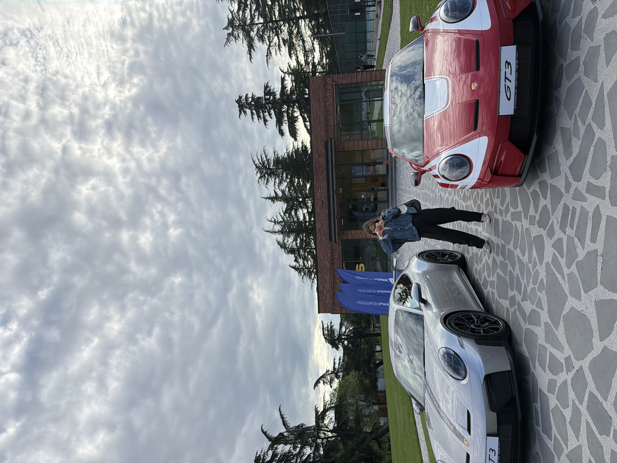 A wide-angle, outdoor photograph depicts a woman standing between two Porsche GT3 cars on a patterned, light-gray paved surface. The upper car is predominantly red with white stripes and the license plate "GT3" is visible. The lower car is a light silver color, with a "PORSCHE" logo visible on its side and a black "GT3" license plate.
The woman is wearing a denim jacket and black pants. Behind the cars, a single-story brick building with large windows and glass doors is visible. To the left, there are three blue flags and coniferous trees.
The sky is overcast with predominantly gray clouds. The image is captured from a low angle and is rotated sideways. The location is identified as Napareuli, Georgia.