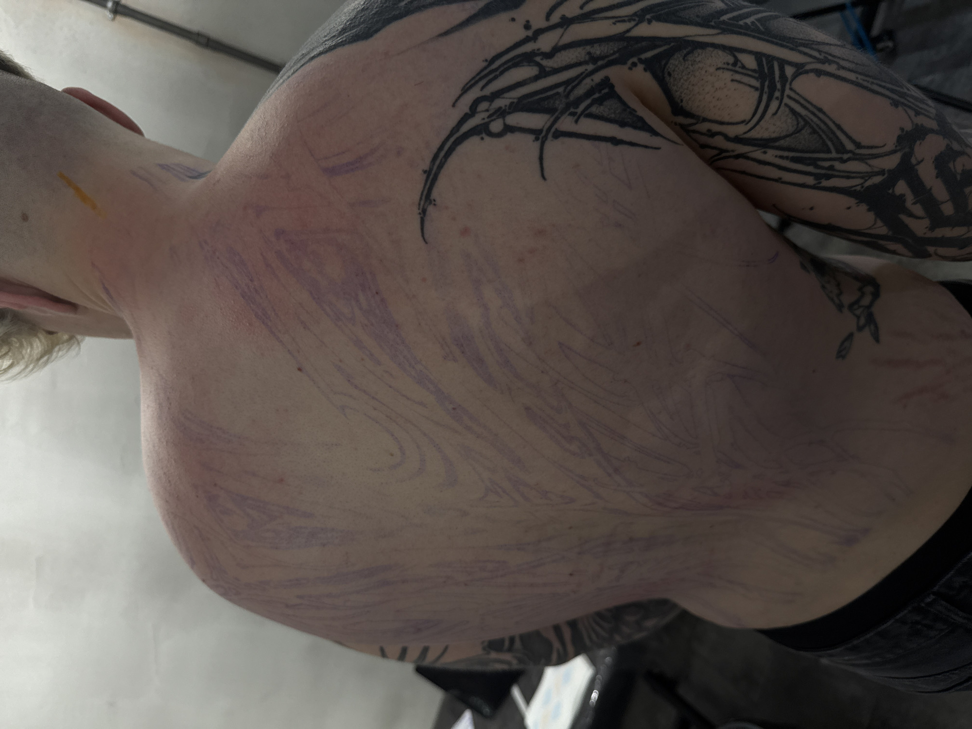 The image shows a close-up of a person's torso and upper arm, featuring multiple tattoos. The left upper arm and shoulder have a detailed, dark-ink tattoo with abstract, organic shapes. The back and side of the torso are covered with a light purple stencil pattern, likely for a new tattoo in progress. The person has short, light-colored hair. The lower portion of the frame shows a glimpse of dark clothing, possibly jeans. The environment appears to be a tattoo studio, suggested by the clinical appearance of the surfaces and the nature of the subjects. There is a yellow stain visible in the left side of the nape. The setting does not provide enough evidence to pinpoint it in Moscow, Russia.