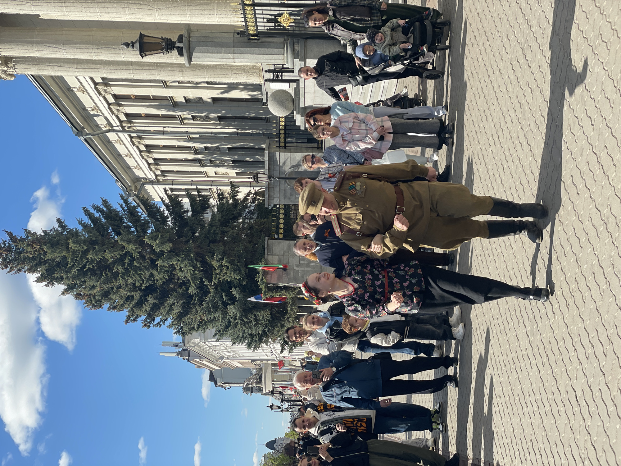 A line of people stands on a paved street in Kazan, Russia, near a building with columns. A person dressed in a military uniform with medals stands out in the line. Flags from different countries, including Bulgaria, are visible. Some people are using phones to take pictures. The sky is blue with clouds, and there is a large tree near the building. A light post is visible on the building. A person with a stroller is in the background.