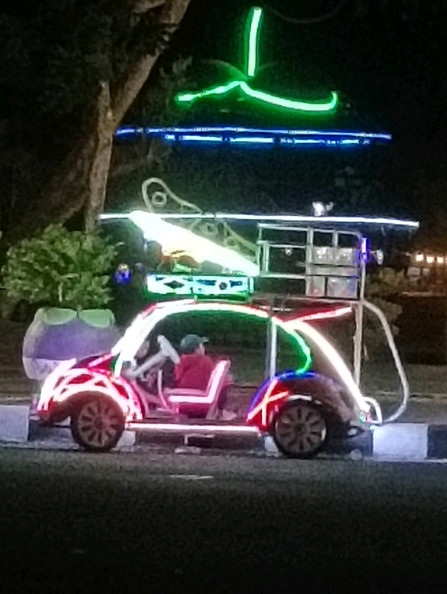 An illuminated vehicle resembling a Volkswagen Beetle is parked on an asphalt surface at night. The vehicle is decorated with neon lights in red, green, and blue. A person wearing a red shirt and a blue cap is seated in the driver's seat. In the background, there are trees, a large purple decorative planter, and building lights. The scene is set in Purworejo, Indonesia, likely a public area given the decorative lighting and the open space. The overall impression is of a festive or decorative display.