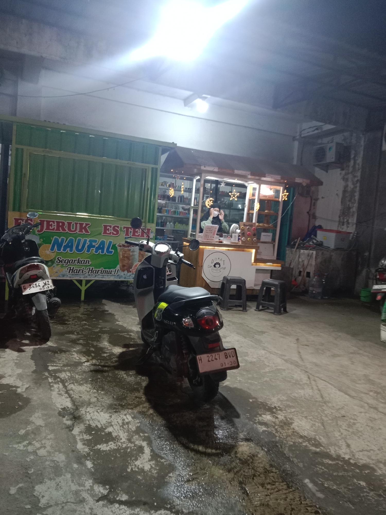 The image depicts an outdoor scene at night, likely in Kebonsari, Indonesia, featuring two parked motorcycles in the foreground and a food stall in the background. The food stall is illuminated and appears to be selling beverages and possibly donuts, as indicated by signage. One motorcycle is a light-colored scooter, and the other is a darker-colored motorcycle. A person wearing a hijab and a mask is visible inside the food stall, possibly serving a customer. The ground is wet, suggesting recent rain or cleaning. The overall atmosphere is somewhat dim, with artificial lighting from the stall and overhead sources.