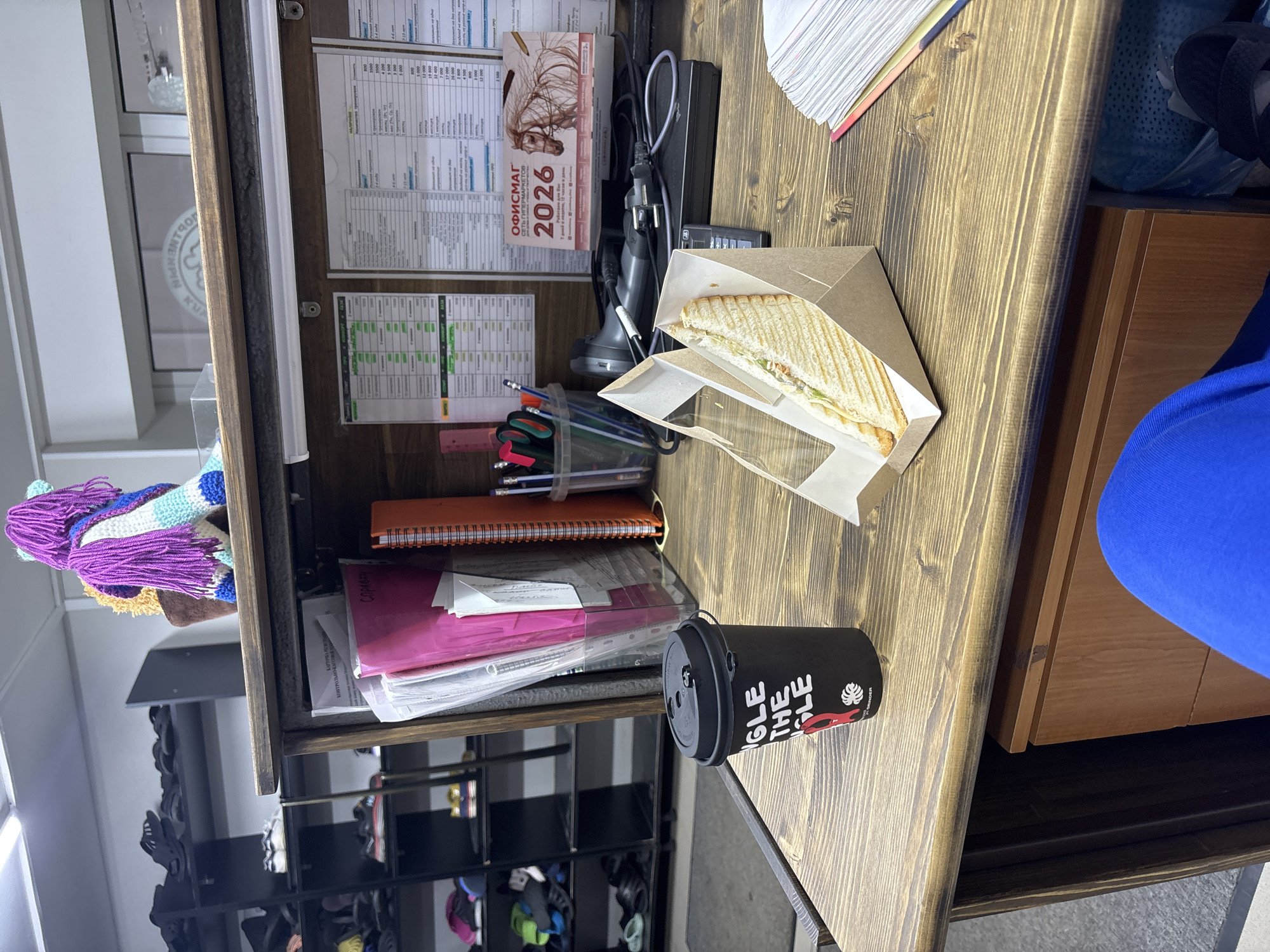 The image shows a desk in Perm, Russia. On the desk are a calendar marked with "2026", a sandwich in a cardboard container, a cup of coffee, a pen holder filled with pens, a notepad, and some paperwork. The desk is made of wood and has a shelf above it. A shoe rack is visible below the desk. A person wearing a blue top is also visible in the image.