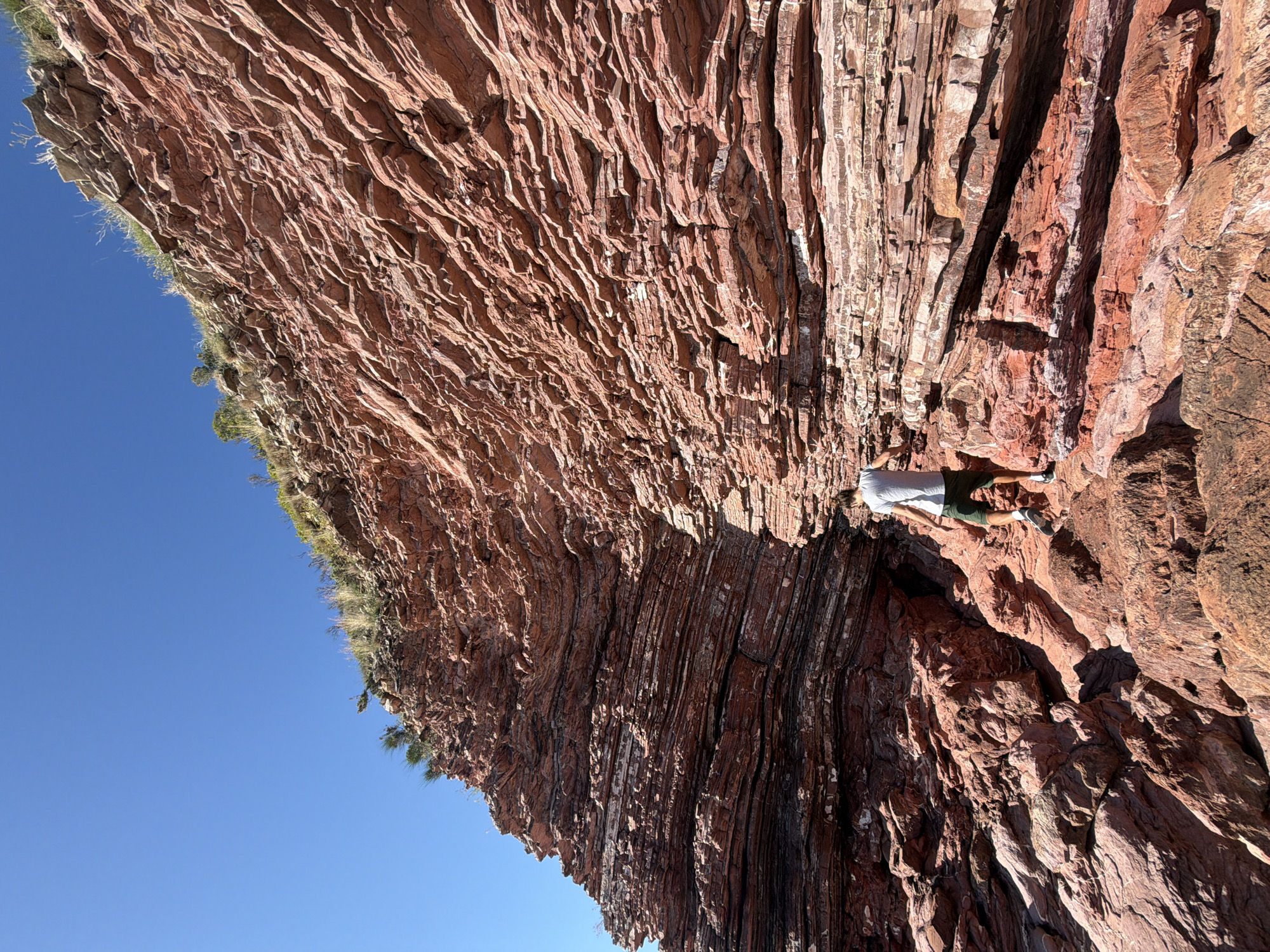 The image shows a cliff face in Sveti Stefan, Montenegro, composed of layers of reddish-brown rock with visible striations. Green vegetation grows along the cliff's edge. A person is positioned on the cliff face, wearing a light-colored shirt and dark shorts, with their feet near a crevice. The sky is clear and blue.
