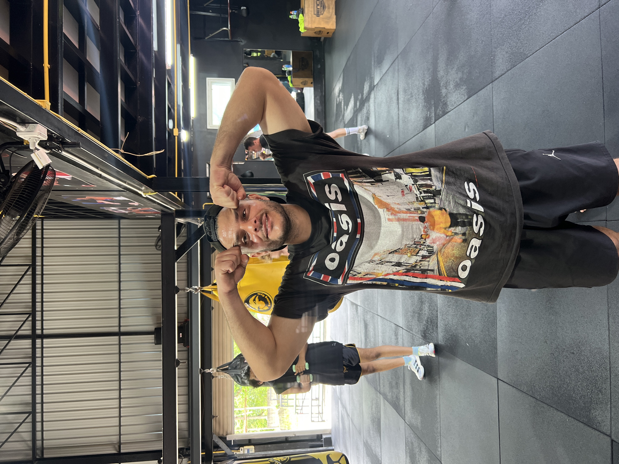 In Chalong, Thailand, a man in a black t-shirt and black shorts flexes his biceps in a gym. The t-shirt features a graphic of a city street scene and the word "Oasis." He wears a black baseball cap. In the background, another person is visible holding two dumbbells. Punching bags hang from the ceiling, and gym equipment is scattered around the room. The gym has a black rubber floor and industrial lighting.