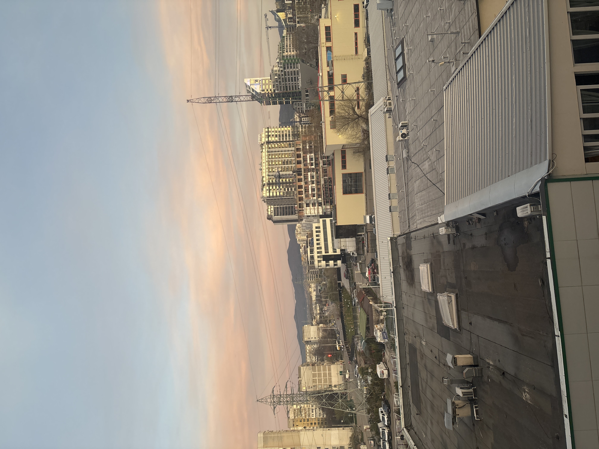 An elevated cityscape scene captured during twilight in Sofia, Bulgaria. The image is oriented in a vertical format. In the middle ground, a visible mountain range sits behind a cluster of high-rise buildings and active construction sites, indicated by tall cranes. The sky is a blend of pale blue and soft orange-pink hues due to the setting sun. Electrical power pylons and their connecting lines dominate the central view, crisscrossing the sky. The foreground consists of rooftops made of metal and dark, tar-like material. Visible air conditioning units and skylights are scattered across the rooftops. The horizon is lined with a variety of buildings, ranging from residential blocks to commercial structures. Car traffic and general city activity are visible.