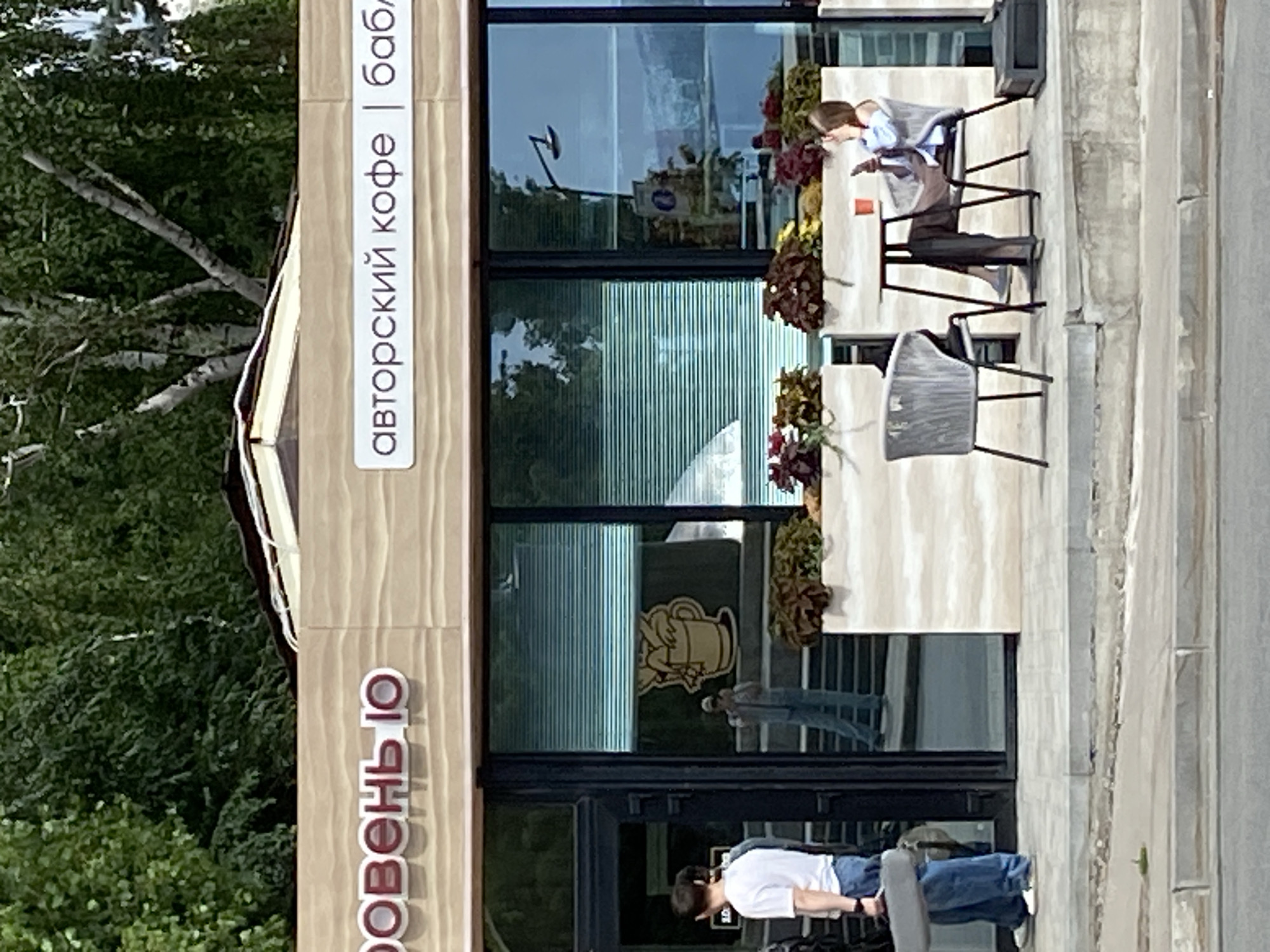 An outdoor cafe scene is depicted in this image. In the foreground, a sidewalk runs horizontally across the bottom of the frame. Behind the sidewalk is a raised concrete patio area. On the patio, two chairs and a small table are visible. A woman is seated at the table, holding a phone. A person in a white t-shirt and blue jeans stands to the left of the image, holding a grey flat object. A building with a large glass storefront is behind the patio. The building's facade has signage that reads "авторский кофе | бабл" and "РОВЕНЬЮ". Green trees are visible in the background, with some branches extending over the top of the building.
