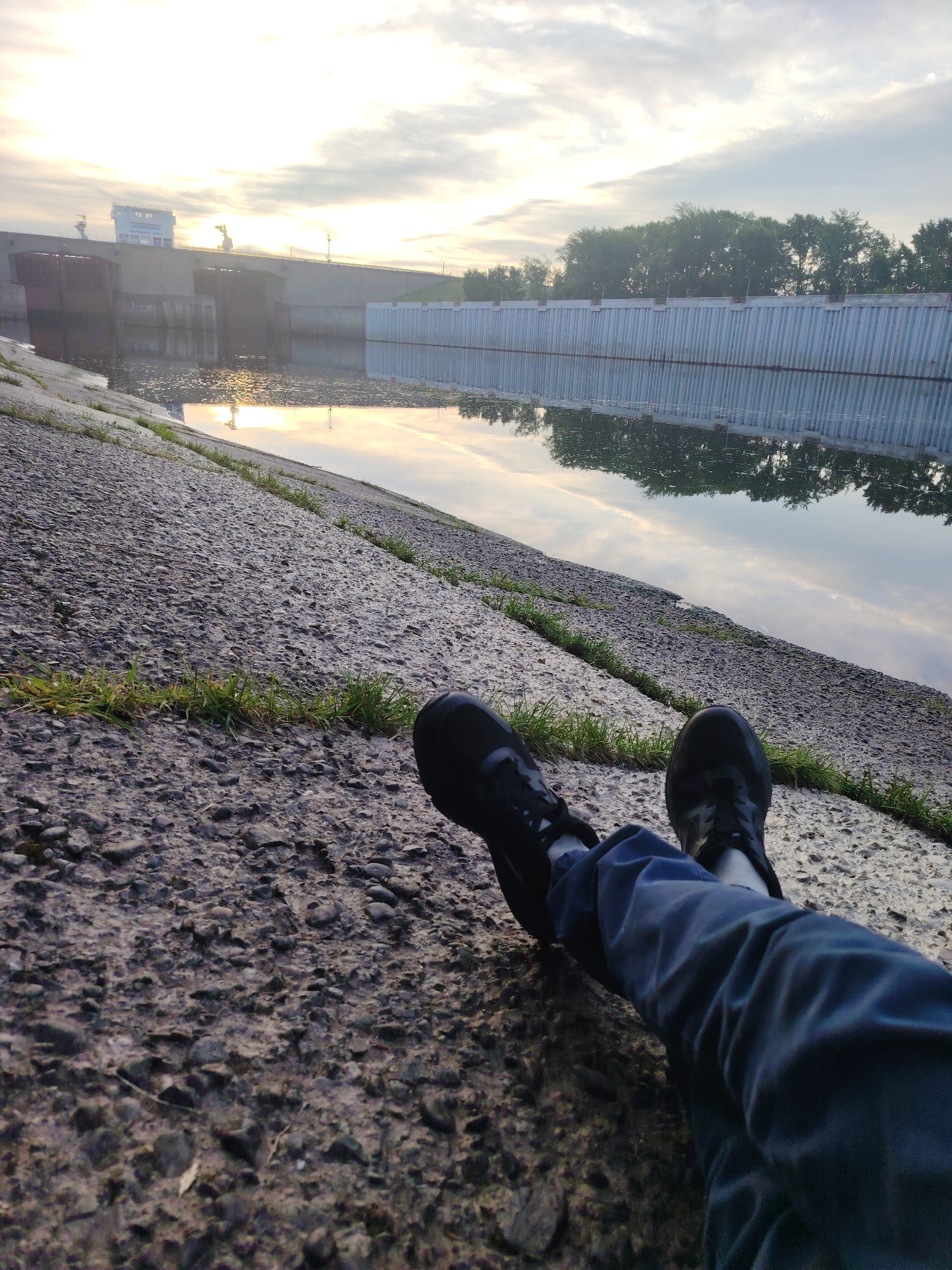 The image presents a first-person perspective from a low angle, showing a person's legs and feet resting on a sloping concrete bank next to a body of water. The person is wearing dark trousers and black sneakers, with their legs extended towards the water. The concrete bank is rough and textured with small pebbles and sparse patches of green grass growing in its cracks.
The calm body of water, likely a river or canal, stretches into the background, reflecting the cloudy sky and the surrounding structures. In the distant left, a large concrete dam or water lock structure is visible, topped by a white building with a blue roof and some industrial elements, possibly cranes. Warm sunlight, characteristic of early morning or late evening, illuminates parts of the scene and creates bright reflections on the water, particularly near the dam.
On the far bank, a tall, corrugated metal wall lines the water's edge, its vertical pattern mirrored clearly in the still water. Beyond this wall, a dense line of green trees extends across the horizon, also reflected in the water's surface. The sky is predominantly overcast with bright, diffused light breaking through. The scene conveys a peaceful and contemplative atmosphere by a man-made waterway, consistent with locations found in areas like Chaikovsky, Russia. No visible text is present in the image.