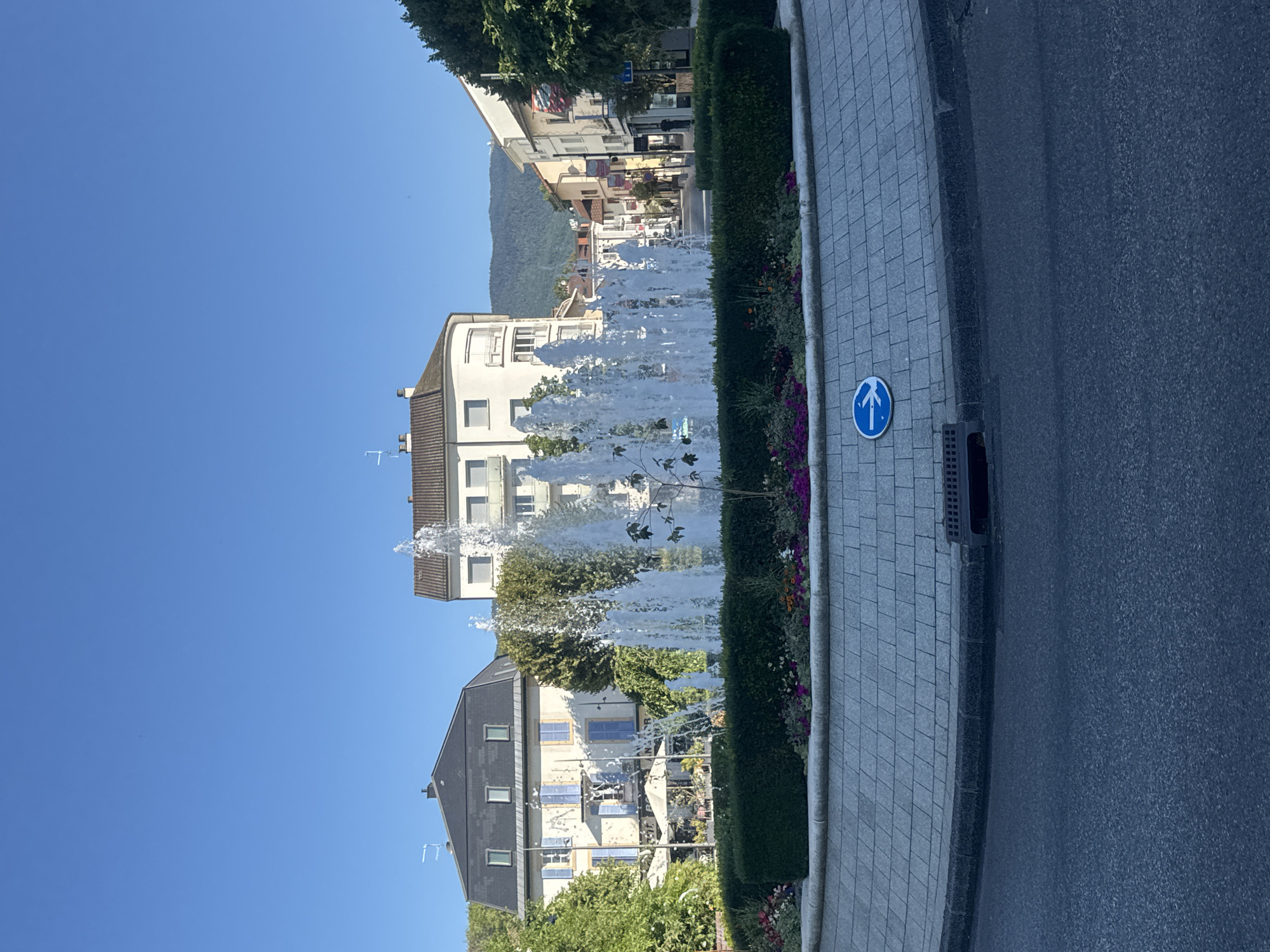 This image shows a public fountain in a roundabout in Divonne-les-Bains, France. The fountain is centrally located, with water jets spraying upwards. Around the fountain is a circular garden bed filled with colorful flowers and green bushes. The roundabout is paved with light-colored interlocking stones, and a blue traffic sign with a white arrow indicating a left turn is visible on the pavement. Beyond the roundabout, the scene depicts a street lined with multi-story buildings, some with balconies. Trees and hills are visible in the background under a clear blue sky. The overall impression is of a pleasant, well-maintained urban square.