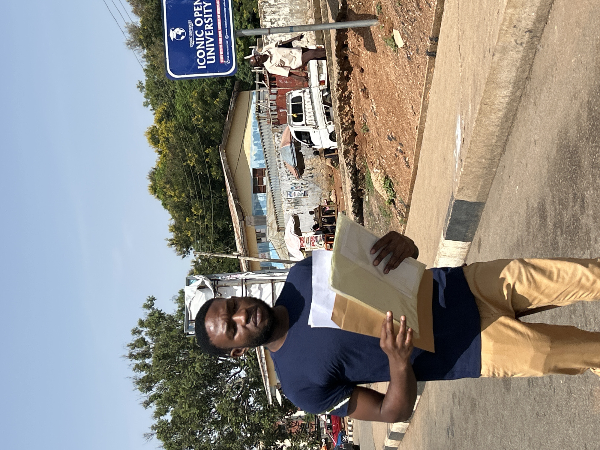 A young Black man wearing a navy blue t-shirt and tan pants walks down a paved road, holding a stack of papers and folders. He appears to be in motion, possibly walking towards or away from a university. In the background, a white truck is parked near buildings that resemble shops or small businesses. A sign for "ICONIC OPEN UNIVERSITY" is visible on the right side of the image, suggesting the location might be near a university campus. The scene takes place outdoors on a sunny day with a clear blue sky. The environment appears to be an urban or semi-urban setting in Jos, Nigeria, characterized by buildings, road infrastructure, and vegetation. Another individual, an older Black person in a light-colored shirt and dark pants, walks further down the road in the background.