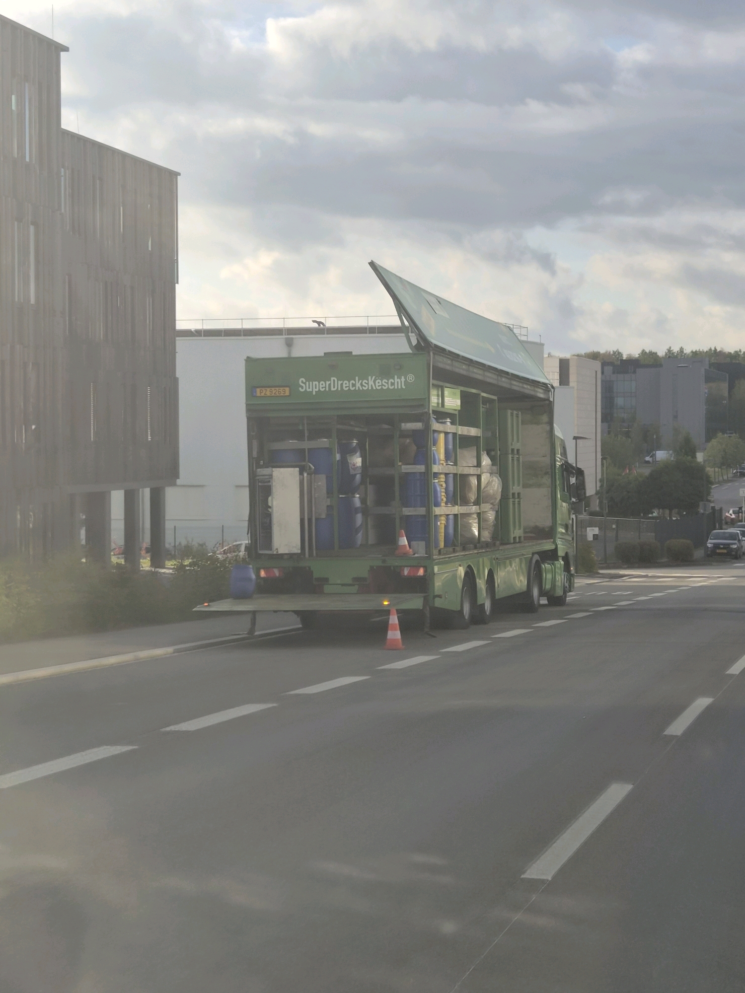 An overcast daytime image shows a green truck with the text "SuperDreckesKëscht" on its side parked on a street in Leudelange, Luxembourg. The truck has an open-top cargo area filled with blue barrels and white bags. An orange traffic cone is positioned on the road near the back of the truck. In the background, there are modern buildings with wooden accents and other commercial structures. A few cars are visible on the road in the distance. The sky is cloudy, suggesting a possibly humid or rainy day.