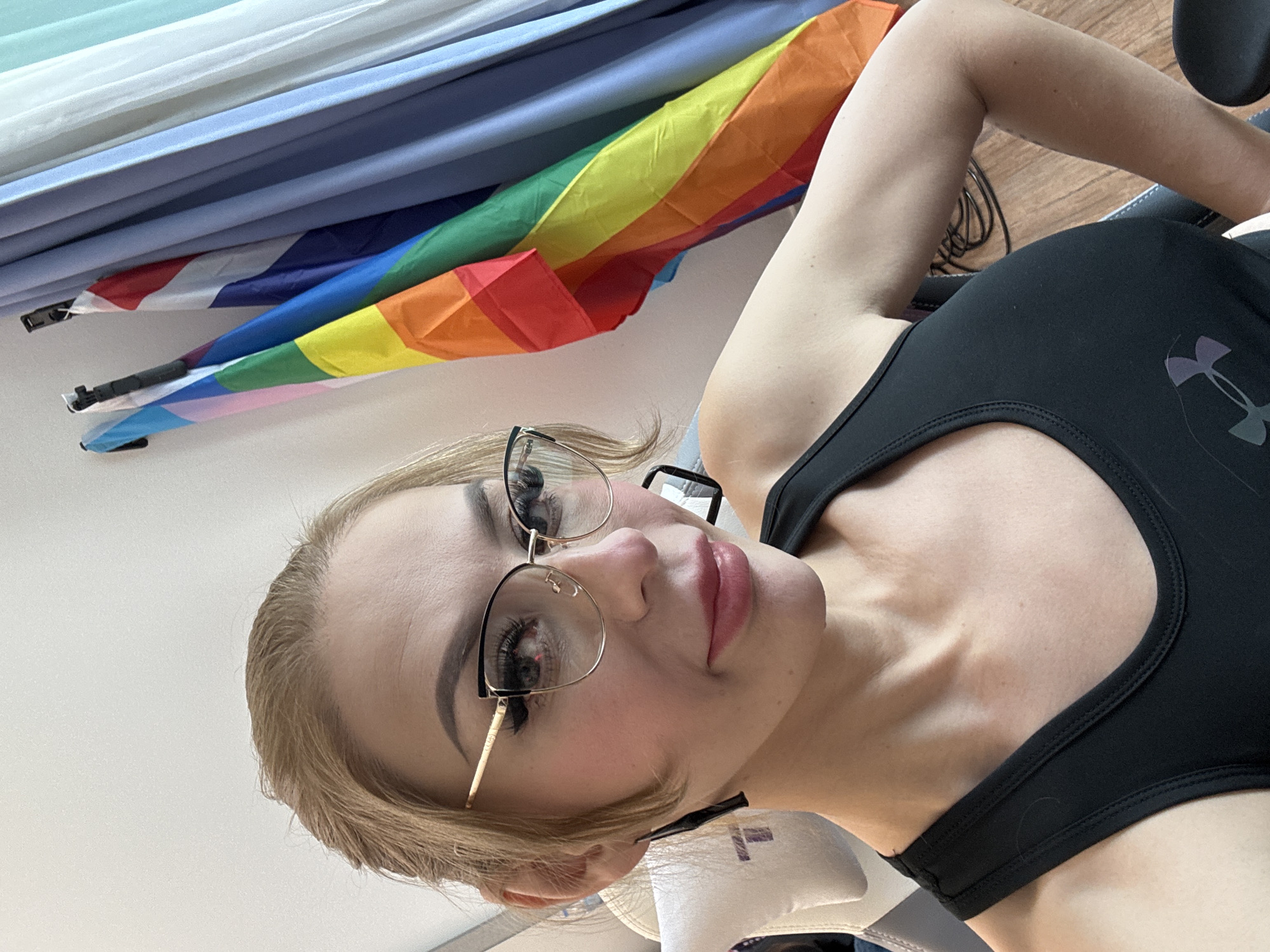 A medium shot photograph shows a woman with blonde hair, wearing glasses, a black tank top, and makeup, posing for a selfie. In the background, several flags are visible, including a rainbow flag and the flag of Thailand. The woman is sitting in a chair, and the environment appears to be indoors, with curtains and a wooden floor. The location is identified as the city of Hua Hin City Municipality, Thailand.