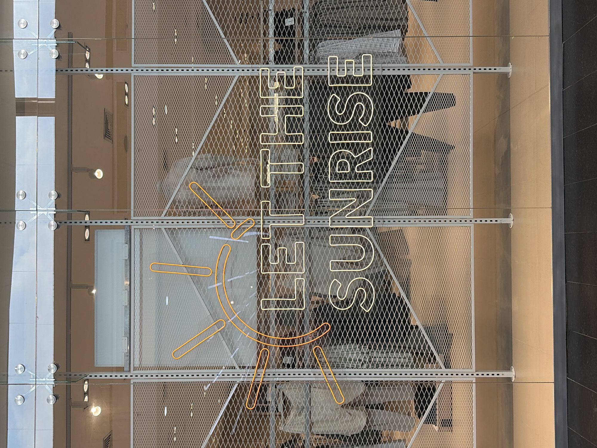 The image captures a static display through a large glass window, likely within a modern commercial or retail setting in Ciudad Guayana, Venezuela. On the left, several large glass panels are visible, secured by circular metal fasteners, reflecting overhead lights and interior elements.
Behind the glass, a prominent light grey metal mesh partition, characterized by a diamond expanded metal pattern, serves as a backdrop for a neon sign. The sign features a stylized sun icon rendered in bright orange/yellow neon, consisting of a central curved line and several radiating lines representing rays. Adjacent to and partially overlapping the sun icon, the phrase "LET THE SUNRISE" is spelled out in bold, white/light yellow block-style neon letters. The words "LET THE" are positioned above "SUNRISE."
The metal mesh is supported by visible horizontal and vertical framing elements. The horizontal frames exhibit a series of small, square perforations. Vertical supports on the right extend downwards to touch a light beige floor, terminating in round bases. Through the mesh and beyond the display, glimpses of an interior space reveal beige walls and various ceiling lights, including circular and square fixtures. To the far right, a darker, possibly tiled wall is visible, marking the edge of the immediate display area. The scene depicts a well-lit, contemporary indoor environment.