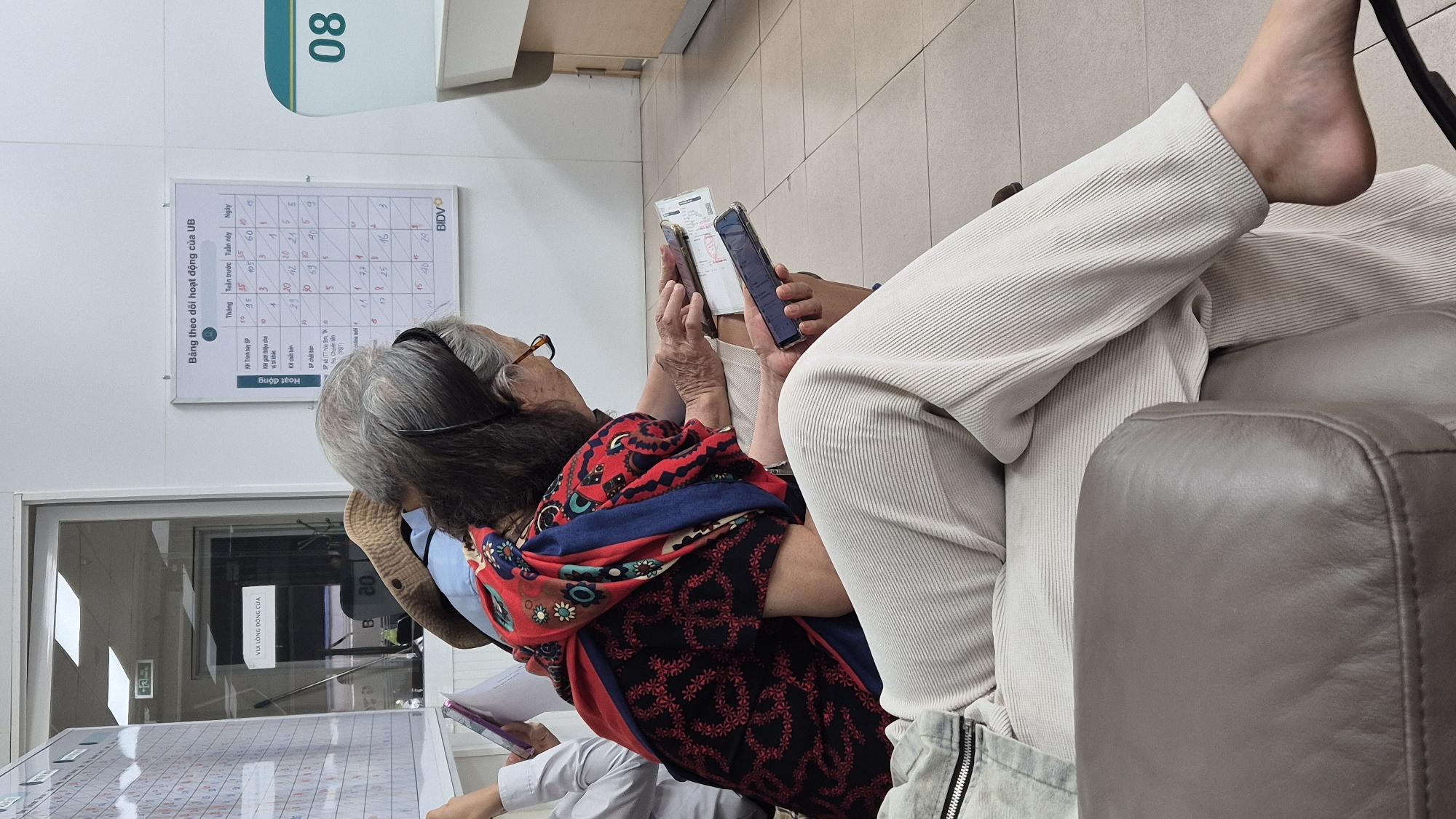 The image depicts an indoor scene, likely a waiting area, with a focus on two individuals. The person in the foreground is wearing light-colored, ribbed pants and a denim jacket. Their left leg is visible, with a bare foot resting near the bottom right of the frame. This individual is seated on a brown leather armchair, only the armrest and a portion of the seat are visible.
The second individual, positioned behind and to the right of the first, is an elderly woman wearing glasses and a vibrant, patterned top with red, blue, and black elements, along with a colorful scarf draped around her shoulders. She is also seated and appears to be looking at a mobile phone she holds in her right hand. Her left hand rests on a document or brochure.
In the background, a whiteboard displays a grid with Vietnamese text and numbers, suggesting a tracking or scheduling system. Text on the whiteboard includes "Bảng theo dõi hoạt động của UB" (UB Activity Monitoring Board), and column headers such as "Tháng" (Month), "Tuần trước" (Last Week), "Tuần này" (This Week), and "Ngày" (Day). A sign on a glass door in the background reads "VUI LÒNG ĐÔNG CỬA" (Please Close Door).
The environment appears to be a public space, possibly a bank or government office, given the presence of the whiteboard and the formal seating arrangement. The overall lighting is neutral, indicating an indoor setting. No specific landmarks or street views are visible to definitively place this scene within the city of Đà Nẵng, Vietnam, however, the Vietnamese text on the signage and whiteboard strongly suggests a Vietnamese location.