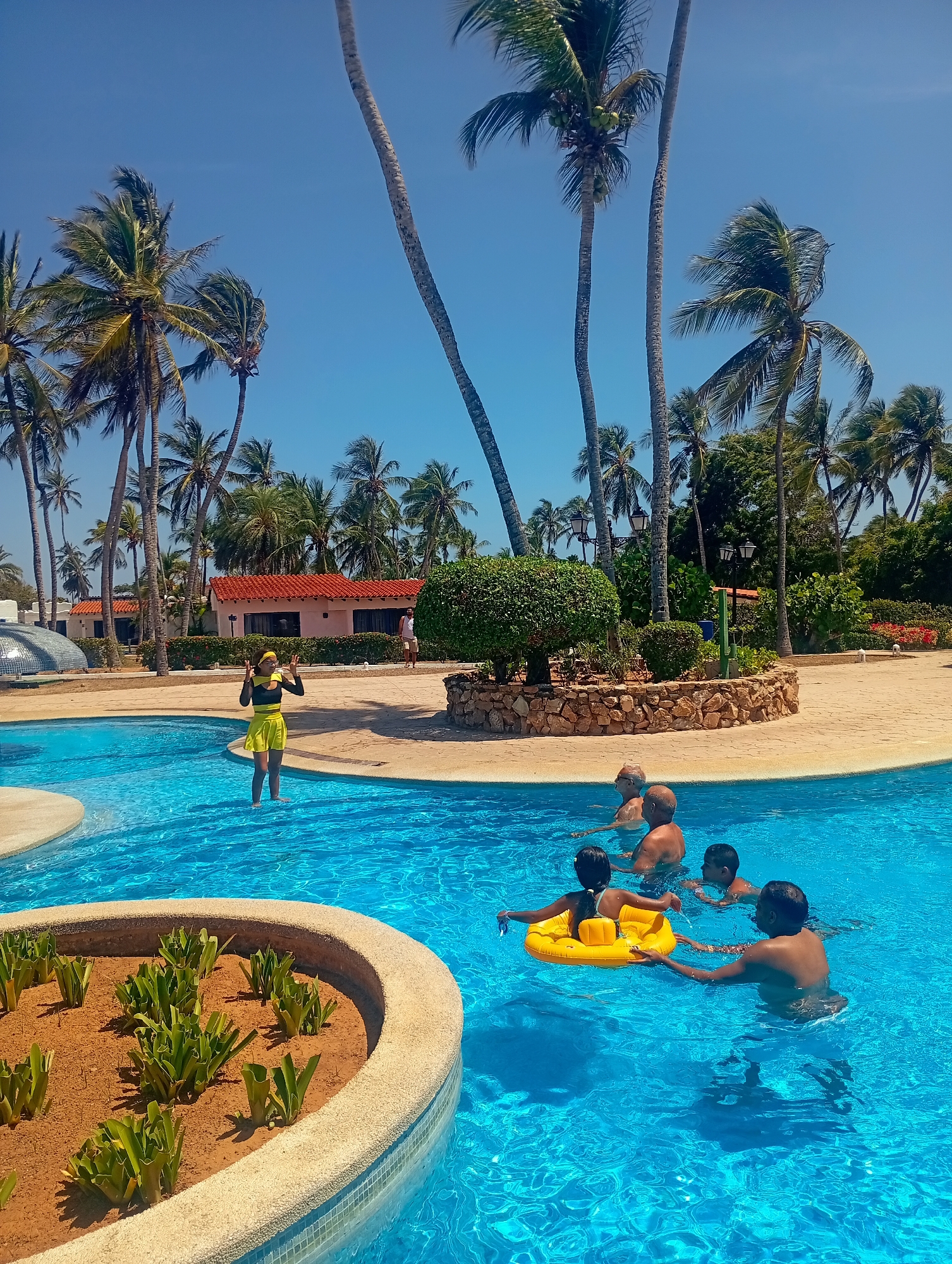 The image depicts a swimming pool scene in what appears to be a resort at Playa El Agua, Venezuela. Several palm trees are in the background. A woman in a yellow and black outfit is standing near the edge of the pool. In the pool, a group of people is swimming, including a child in a yellow inflatable ring. One adult appears to be helping the child. There is a small raised garden with plants in the pool. In the background, there are buildings with red tile roofs and some landscaping. The sky is blue and clear.