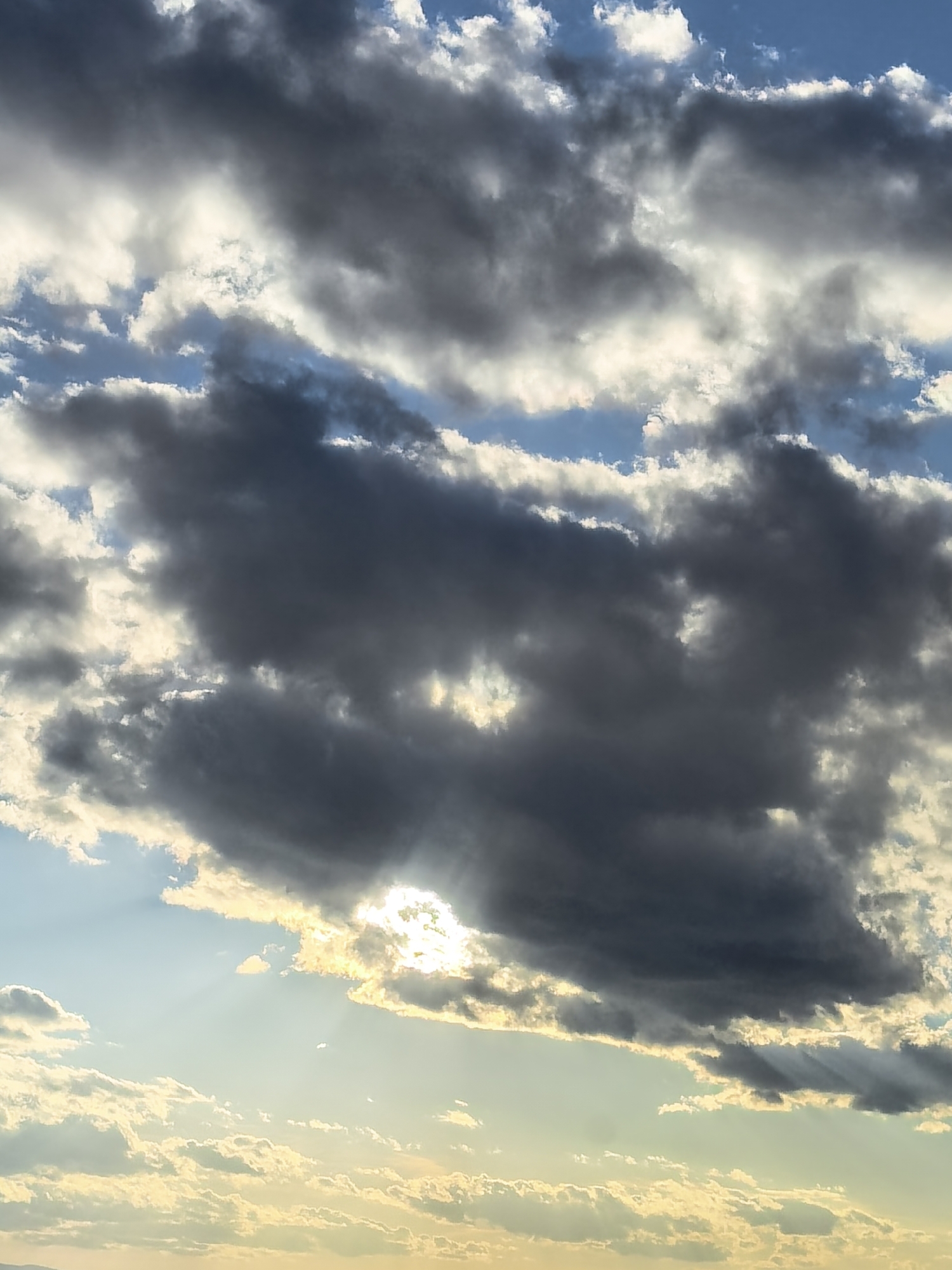 The image captures a dramatic sky with a large, dark cloud dominating the view. Sunlight streams through breaks in the cloud cover, creating a striking contrast between light and shadow. The upper portion of the image reveals patches of blue sky interspersed with white clouds, while the lower section is filled with lighter, golden-hued clouds closer to the horizon. No people or specific objects are visible in the environment. The location is Unknown, Unknown.