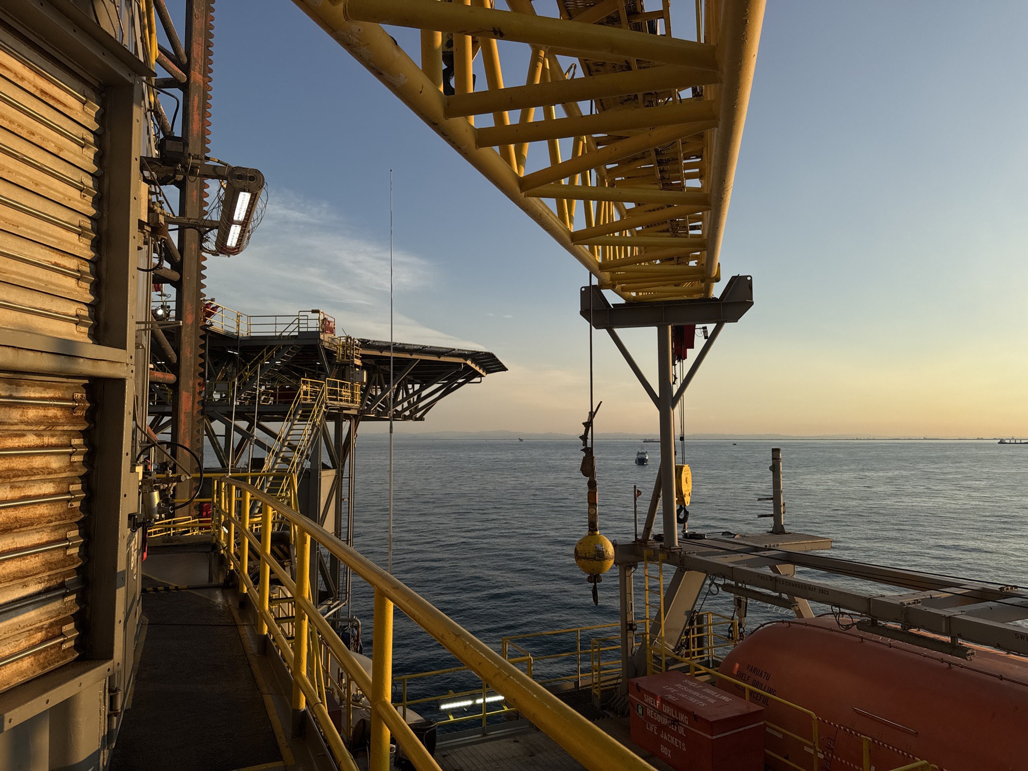 The image depicts an industrial scene on an offshore oil rig under a clear sky at sunset. A large yellow crane boom extends across the top of the frame. The foreground features a metal walkway with yellow safety railings. To the left, a corrugated metal wall is visible. Further back, metal structures, stairs, and safety equipment like life jackets in red boxes are present. The sea stretches out to the horizon, with several ships and what appear to be distant land structures or ports visible. The lighting suggests it is either sunrise or sunset. There are no people directly visible in the frame.