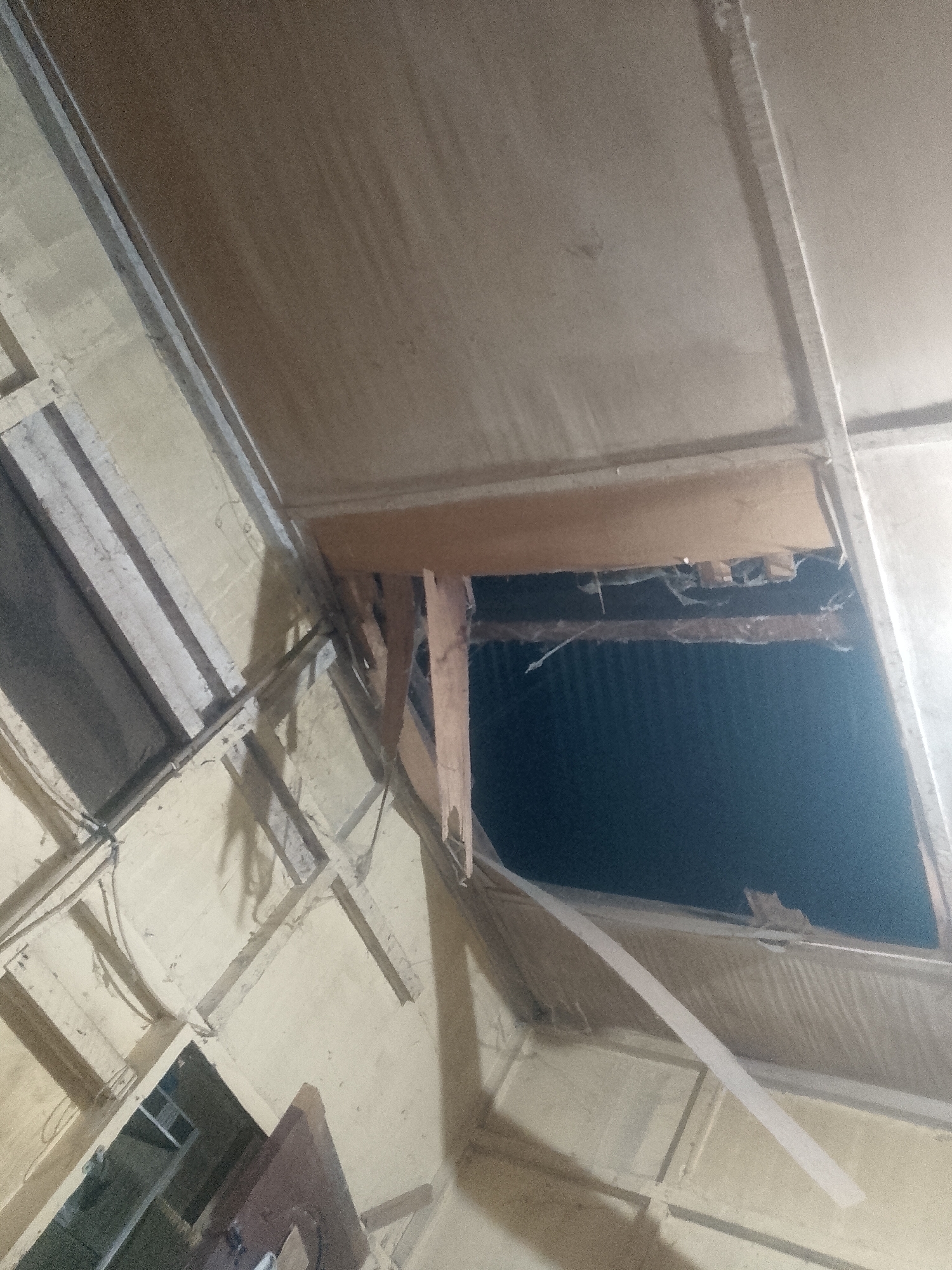 The image shows a damaged interior ceiling with a large hole exposing the roof structure and potentially the exterior. The ceiling is made of wooden panels and beams, and portions are broken or missing. The darkness visible through the hole suggests nighttime or a poorly lit exterior. Given the mention of Lhokseumawe, Indonesia, it's likely the building is a residential or commercial structure impacted by weather or structural issues common in the region. No people are visible. The only identifiable objects are parts of the building's damaged ceiling and roof. There is no visible action or interaction beyond the overall state of disrepair. The environment appears enclosed, but the damaged ceiling exposes it to the elements.