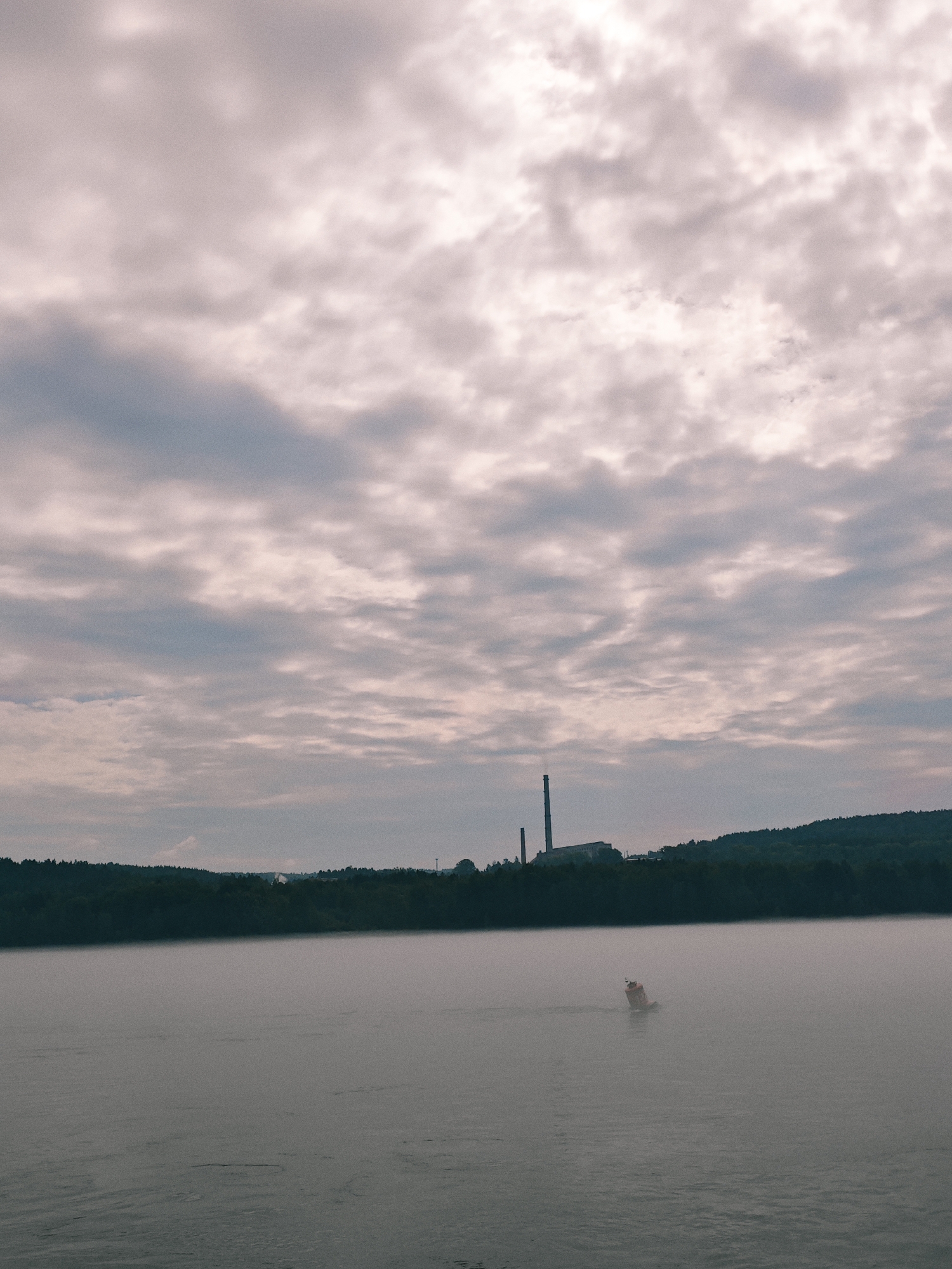 The image depicts a hazy, overcast sky over a wide body of water. In the distance, a dark, tree-covered shoreline is visible, with industrial structures, including a tall smokestack, situated on the horizon. A small, orange buoy is floating in the misty water, positioned off-center to the right. The overall atmosphere is subdued and somewhat industrial, characteristic of a waterfront area. The location is identified as Atamanovo, Russia.