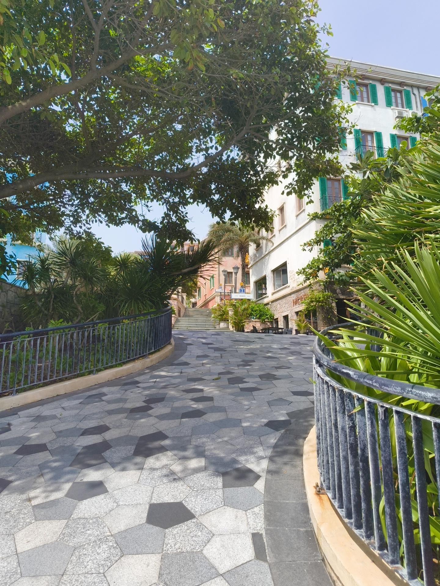 The image captures a paved walkway with a geometric hexagonal pattern, curving to the right. The walkway is bordered by a dark metal railing and lush green foliage, including palm-like plants and large trees with dense leaves. In the background, a multi-story building with white walls and green shutters is visible, suggesting a Mediterranean architectural style. Further down the walkway, stairs lead upwards, flanked by more vegetation and buildings with colorful facades, including a bright blue one on the left and an orange one on the right. A street lamp is also visible near the stairs. The scene appears to be a well-maintained public area in a warm climate, possibly a resort town or a coastal city. Based on the context, this location is the Starbay residential area in Phu Quoc, Vietnam.