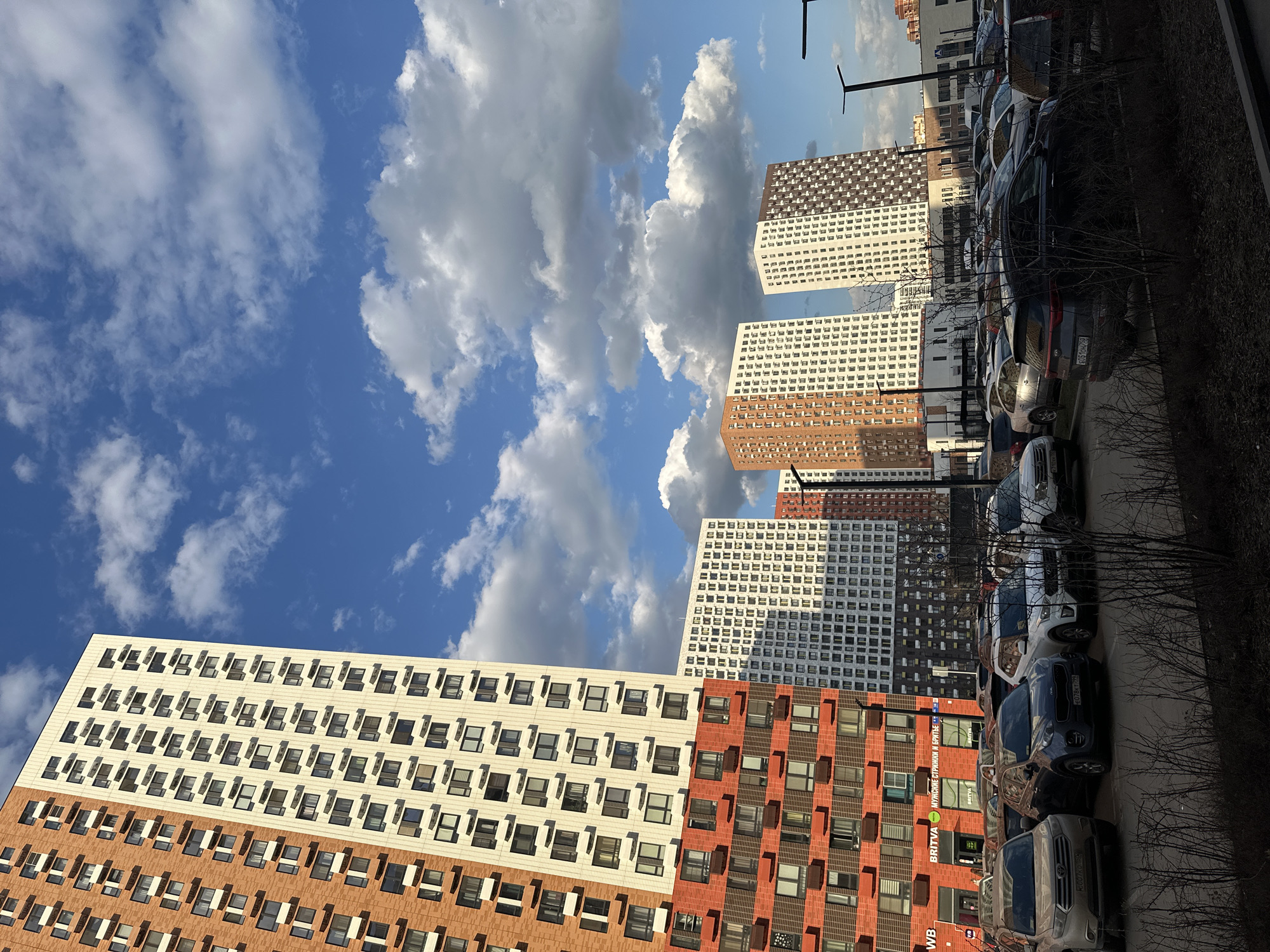 The image shows a view in Lyubertsy, Russia, dominated by modern architecture under a sky filled with cumulus clouds. Several high-rise residential buildings are visible, characterized by alternating bands of white and orange/brown facades with regularly spaced windows. A row of parked cars lines the right side of the frame, partially obscuring the base of the buildings. Bare tree branches intermingle with the cars, suggesting early spring or late autumn. Street lamps are placed intermittently. The lighting suggests it is daytime, with shadows cast by the clouds and buildings. A street sign advertising "BRITVA" which appears to be a barber shop is visible.