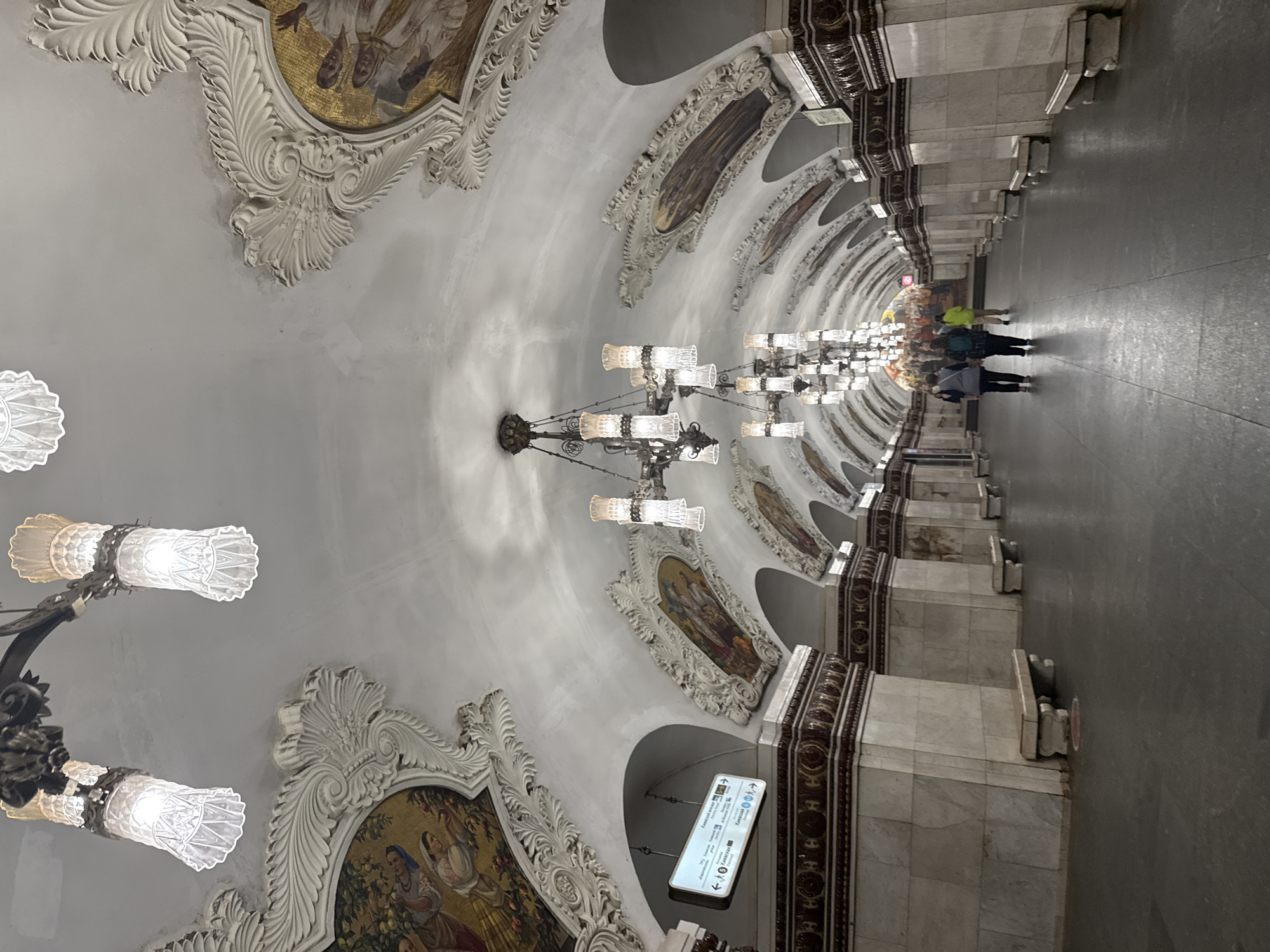 The image depicts a grand, ornate metro station ceiling in Moscow, Russia. The station features an arched ceiling adorned with elaborate white stucco moldings and large, decorative mosaics set within these moldings. Several large, multi-tiered chandeliers with glass elements hang from the ceiling, providing illumination. The station walls are lined with marble columns and a decorative band above the marble. Benches made of marble are positioned along the walls. A directional sign with Cyrillic text is visible hanging from the ceiling on the left side of the frame. In the distance, several people are walking down the platform. The overall impression is one of opulence and historical grandeur, characteristic of some of Moscow's historic metro stations.
