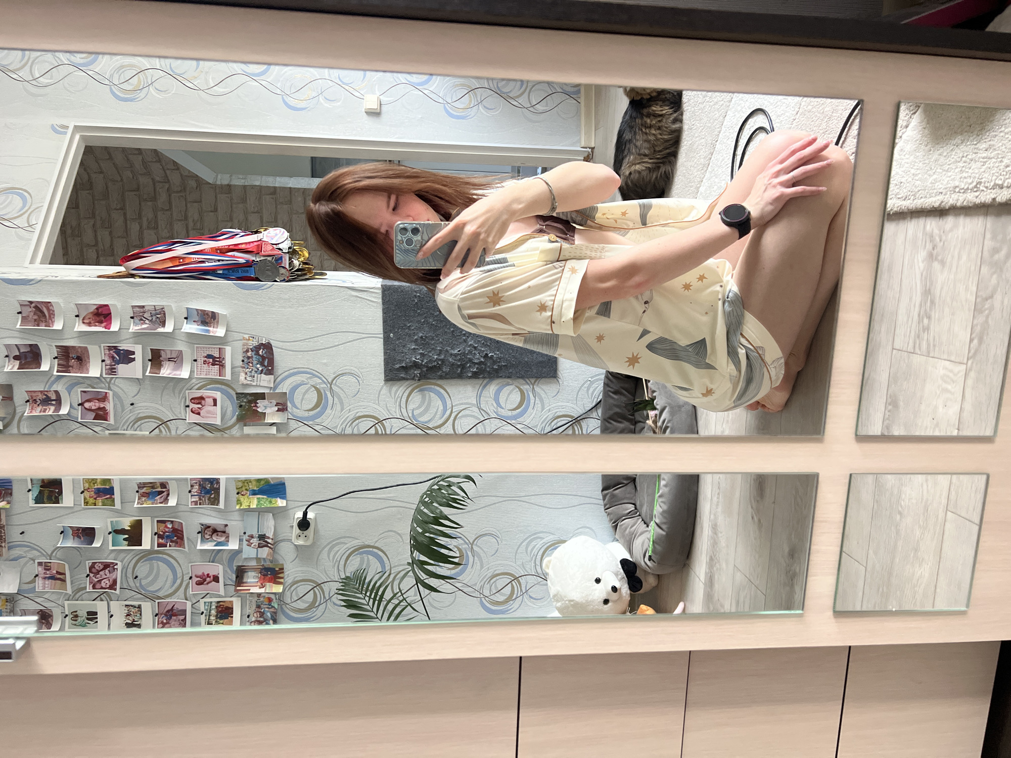 The image displays a mirror selfie taken indoors by a young woman in what appears to be a residential setting in Chaikovsky, Russia. The woman, with long brown hair, is partially visible, seen from her chest to her mid-thighs. She is squatting or kneeling, wearing a light-colored, short-sleeved shirt patterned with stars and abstract natural elements, resembling pajamas. She holds a silver smartphone to capture her reflection, largely obscuring her face. A black smartwatch is worn on her left wrist, and a silver bracelet on her right.
The reflected environment reveals a personalized space. The walls feature light blue and gray swirling patterned wallpaper. On a shelf or ledge below the mirror, numerous small photographs are tacked, showcasing various people and possibly outdoor scenes. Above these photos, a collection of medals with red, white, and blue ribbons is displayed, one of which clearly reads "BIG KVCE," indicating achievements, possibly in sports or academics.
Other reflected details include a fluffy brown tabby cat, likely a Maine Coon, partially visible behind the woman. A white plush toy with a black bow tie and a fern-like houseplant are seen in the lower part of the reflection. The flooring is light-colored wood, partially covered by a white rug. A dark, textured piece of wall art and an opening with a brick-like interior texture are also reflected. The scene portrays a casual, personal moment of self-documentation within her home during what appears to be daytime.