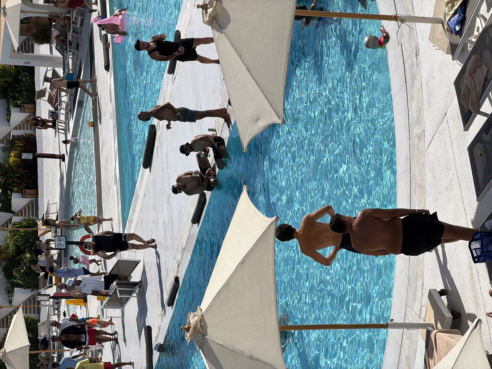 This image depicts a lively poolside scene at a resort, likely in Sharm El Sheikh, Egypt, given the architectural style and abundant sunlight. Multiple people are visible in and around a bright blue swimming pool.
In the foreground, two large, light-colored umbrellas provide shade over lounge chairs. A young man, shirtless and wearing black swim trunks, stands facing away from the camera, his hands on his hips. Another man, also shirtless with dark swim trunks, is positioned behind him, facing slightly left.
Further back, several individuals are in the pool. One man, wearing a black tank top and black shorts, is standing near the edge of the pool, looking towards the right. Another man, in a black tank top and black shorts, is sitting on the edge of the pool, facing left and holding a drink. A third man, shirtless with gray shorts, is also sitting on the edge, smoking a cigarette.
In the middle ground, a group of people are gathered near the pool. A man in a black tank top and black shorts is walking away from the camera. Another man, shirtless with gray shorts, is walking past him. A man in a black t-shirt and black shorts is carrying lounge chairs. A woman in a black bikini stands by the pool, observing. Children are also present, with one girl in a yellow bikini in the water. A pink flamingo float is visible in the pool, with a person in it.
The environment is characterized by a large, sparkling blue swimming pool with white tiling around its edges. The area surrounding the pool is paved with light-colored concrete. Several lounge chairs, umbrellas, and poolside furniture are scattered throughout the scene, suggesting a resort or hotel setting. The overall impression is one of a relaxed, recreational atmosphere on a sunny day.