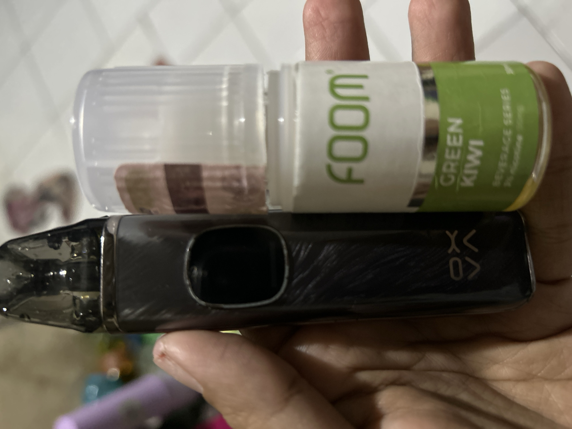 A hand is holding a dark grey vape pen and a bottle of "FOOM GREEN KIWI BEVERAGE SERIES 3% nicotine 30mg" e-liquid. The vape pen has a metallic sheen and a black mouthpiece. The e-liquid bottle is clear with a white cap and a green label. The background is blurred and appears to be an indoor setting, possibly a home or shop, with various out-of-focus objects in shades of pink, blue, and white. The lighting suggests it is daytime. The image's focus is on the hand and the vaping products. The specific location in Depok, Indonesia, cannot be determined from the visual information provided.