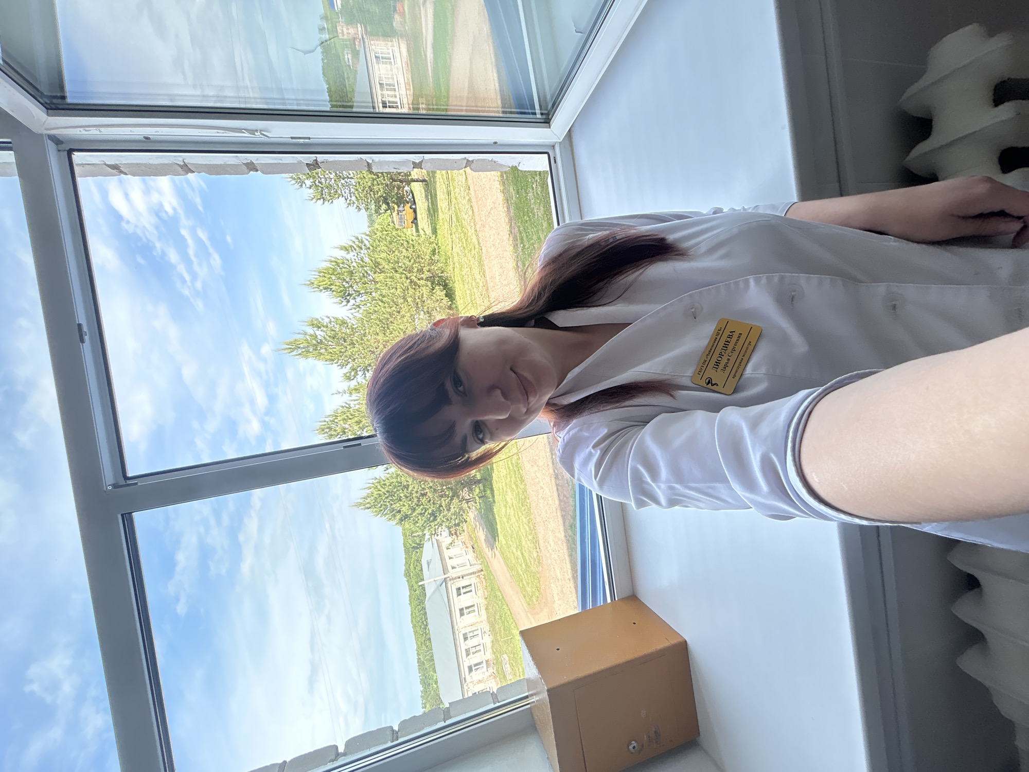 A young woman with dark hair and bangs is visible in the image. She is wearing a white medical coat with a yellow name tag attached to it. She is positioned in front of a window that offers a view of a sunny outdoor scene. The view from the window features a building with multiple windows and a green lawn. There are trees in the background and a blue sky with scattered clouds overhead. In the foreground, there is a metal safe on the windowsill. To the right, a white radiator is visible. The scene appears to be inside an office or similar space. Based on the name tag, the location is assumed to be in Фоки, Russia.