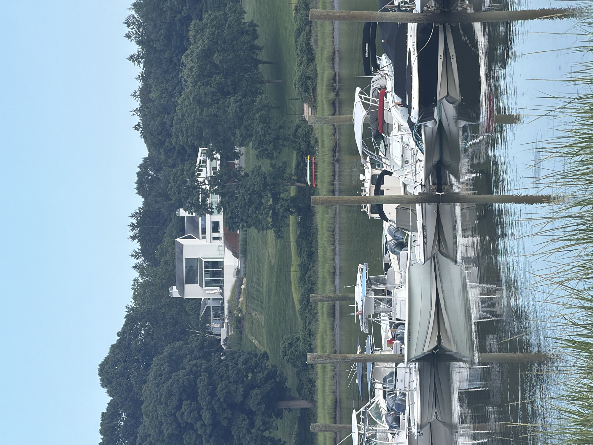 The image shows a body of water with several boats docked. In the background, there is a large modern house on a hill surrounded by trees. The sky is clear and light blue. The boats are of various sizes and types, with some covered and others open. The water is calm, with reflections of the boats and the sky visible. The environment appears to be a marina or a waterfront residential area. The location is identified as the Village of Bayville, United States of America.