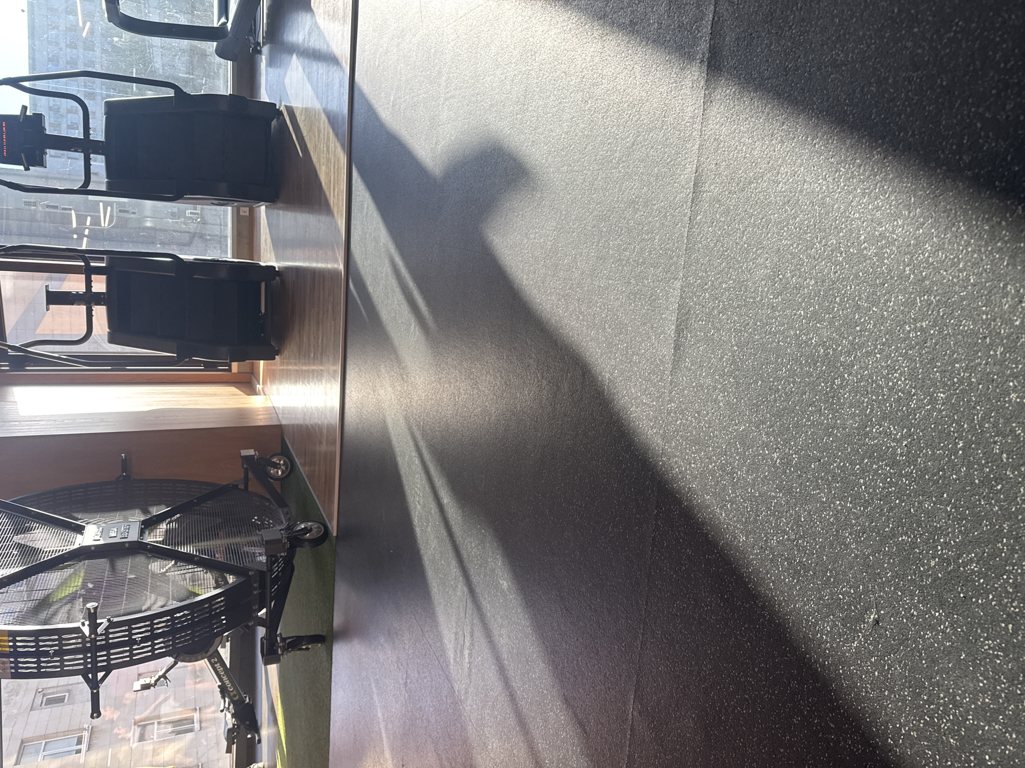 The image captures a bright, sunlit view of a gym or fitness facility in Plovdiv, Bulgaria, focusing on exercise equipment and the floor. The scene is illuminated by strong natural light streaming through large windows, creating prominent, long shadows that diagonally cut across the floor.
On the left side of the frame, several pieces of black exercise equipment are visible, positioned near the windows. These include two vertical machines, identifiable as Concept 2 SkiErgs, with digital monitors at the top. Below them, partially visible, is a large black fan bike, a Concept 2 BikeErg, with the "Concept 2" logo clearly legible on its frame. The machines are placed on a section of light brown, wood-patterned flooring, which contrasts with the majority of the floor space.
The dominant flooring type is a dark, speckled rubber matting, flecked with white or light-colored particles, designed for gym use. The strong sunlight highlights the texture of this matting and casts sharp, elongated shadows from the equipment and window frames onto its surface. Through the windows, a blurred view of exterior buildings, possibly apartments or offices, can be seen under a bright sky.
No people are present, indicating a quiet moment in the gym. The time of day appears to be either morning or late afternoon, given the low angle of the sun suggested by the long shadows. The overall impression is one of a clean, well-lit fitness space.