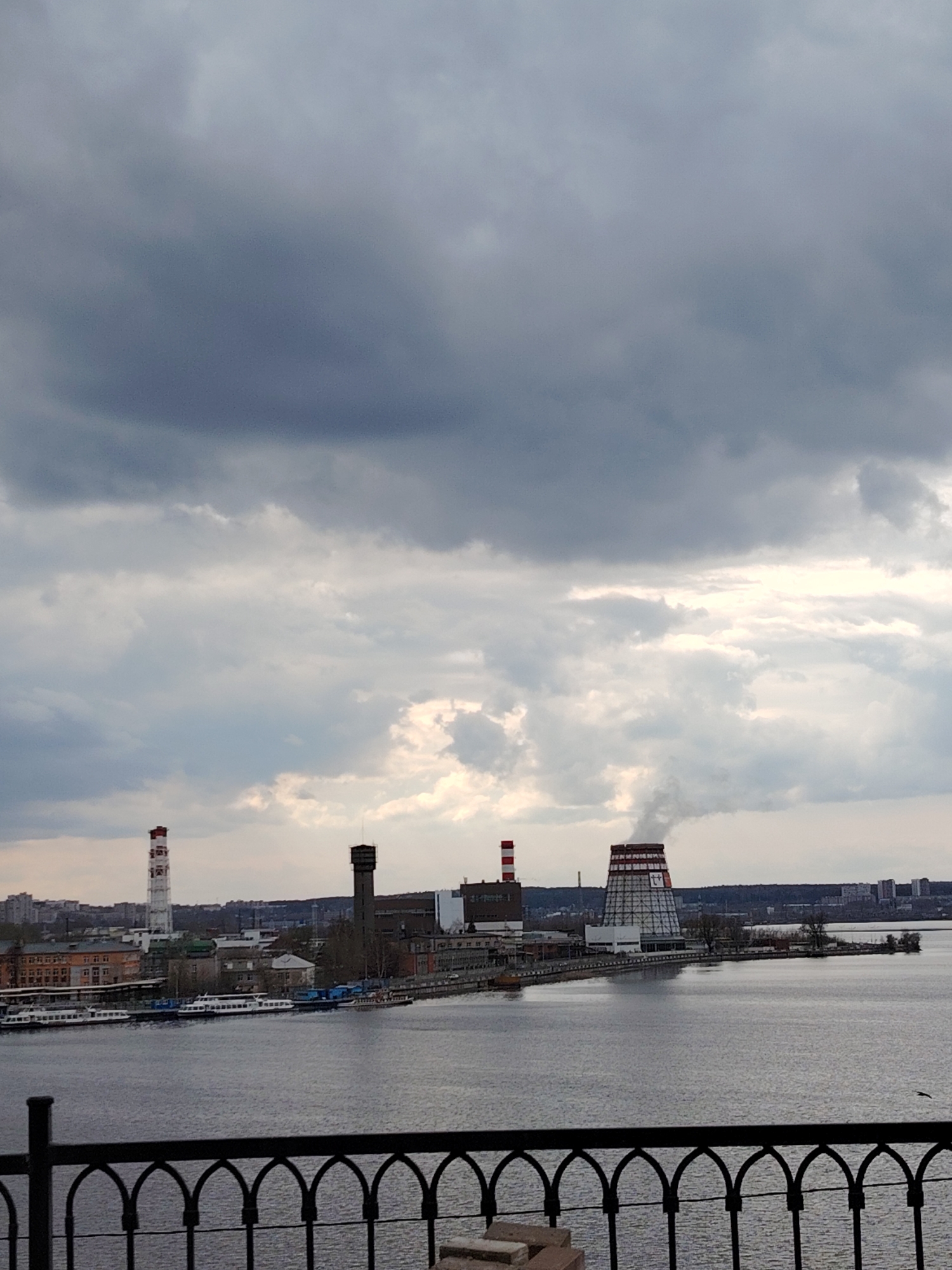 The image captures a view of the Izhevsk Thermal Power Plant-1 (TETs-1) in Izhevsk, Russia, as seen from a slightly elevated perspective. In the foreground is a section of a black wrought-iron fence. Beyond the fence, the foreground transitions into the water of the Izhevsk Reservoir. Several boats are docked along the shoreline near the power plant complex.
The TETs-1 facility itself is a prominent feature of the image, consisting of various industrial buildings and tall structures, including chimneys with red and white stripes, as well as a cooling tower. The power plant sits on a peninsula extending into the reservoir.
In the distance, across the reservoir, the cityscape of Izhevsk is visible, marked by residential buildings and other structures. The sky is overcast with heavy, gray clouds, which dominate the upper portion of the image.