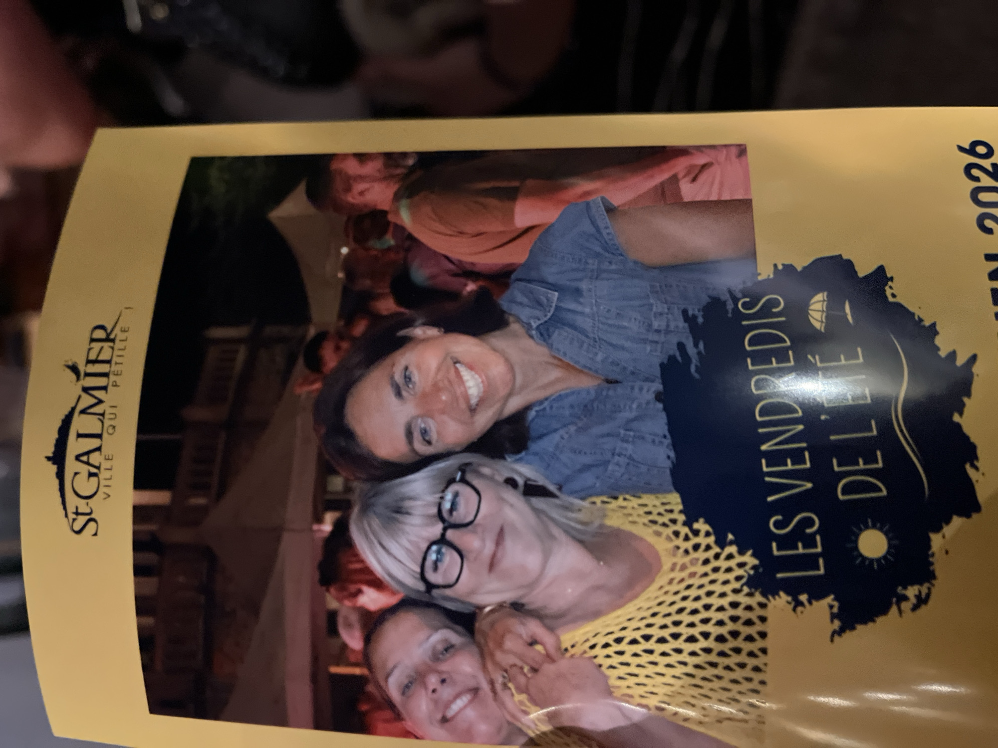 The image shows a photograph of three smiling women at an outdoor event. The woman on the left, wearing a yellow top, is looking at the camera. The woman in the center, wearing glasses and a yellow top, is also smiling at the camera. The woman on the right, wearing a denim shirt, is smiling broadly at the camera. In the background, there are tents and other people enjoying the event. The text on the bottom of the image reads "LES VENDREDIS DE L'ÉTÉ" which translates to "Summer Fridays." Above the photograph, the text "St GALMIER" is visible, indicating that the event is taking place in Saint-Galmier, France. The text "VILLE QUI PETILLE" translates to "Sparkling City." The bottom of the image also shows the year "2026".