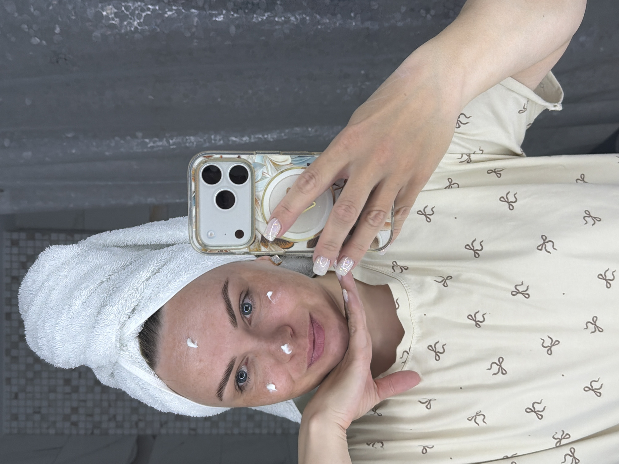 A woman with a towel wrapped around her head is taking a selfie in what appears to be a bathroom. She has dots of white cream on her face. She is holding her phone, which has a decorative case, and posing with her hands near her face. She is wearing a light-colored shirt patterned with brown bows. The background includes what seems to be a shower area and tiled flooring. There is no information in the image to suggest that it was taken in Votkinsk, Russia.