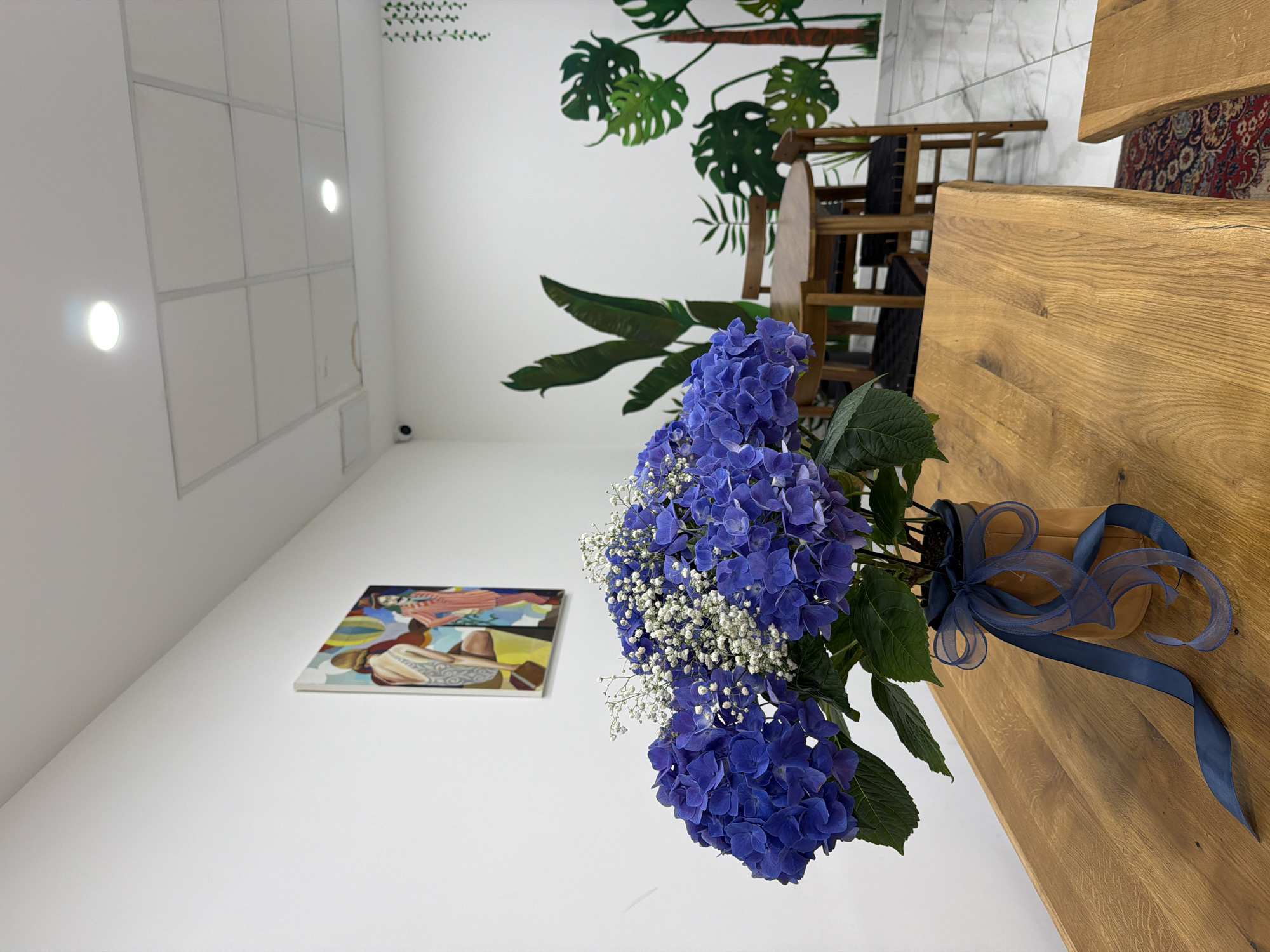 Indoor scene in Sofia, Bulgaria. A bouquet of blue hydrangeas and baby's breath is the focal point, placed on a light-colored wooden table. The bouquet is tied with a blue ribbon. The backdrop includes a white wall adorned with a painting and decorative plant motifs. The ceiling is white with grid-patterned panels and recessed lighting. In the background, a wooden chair and a glimpse of patterned flooring can be seen.