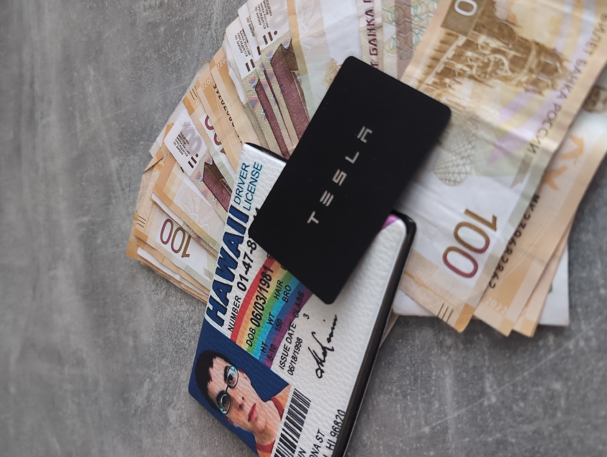 The image depicts a grey surface with various items scattered on it. There's a Hawaii driver's license with a photo of a person with glasses, a black Tesla card, and a stack of Russian rubles. The rubles appear to be 100-ruble notes. The driver's license indicates a birthdate of June 3, 1981. The image does not provide information that can identify the location of Izhevsk, Russia.