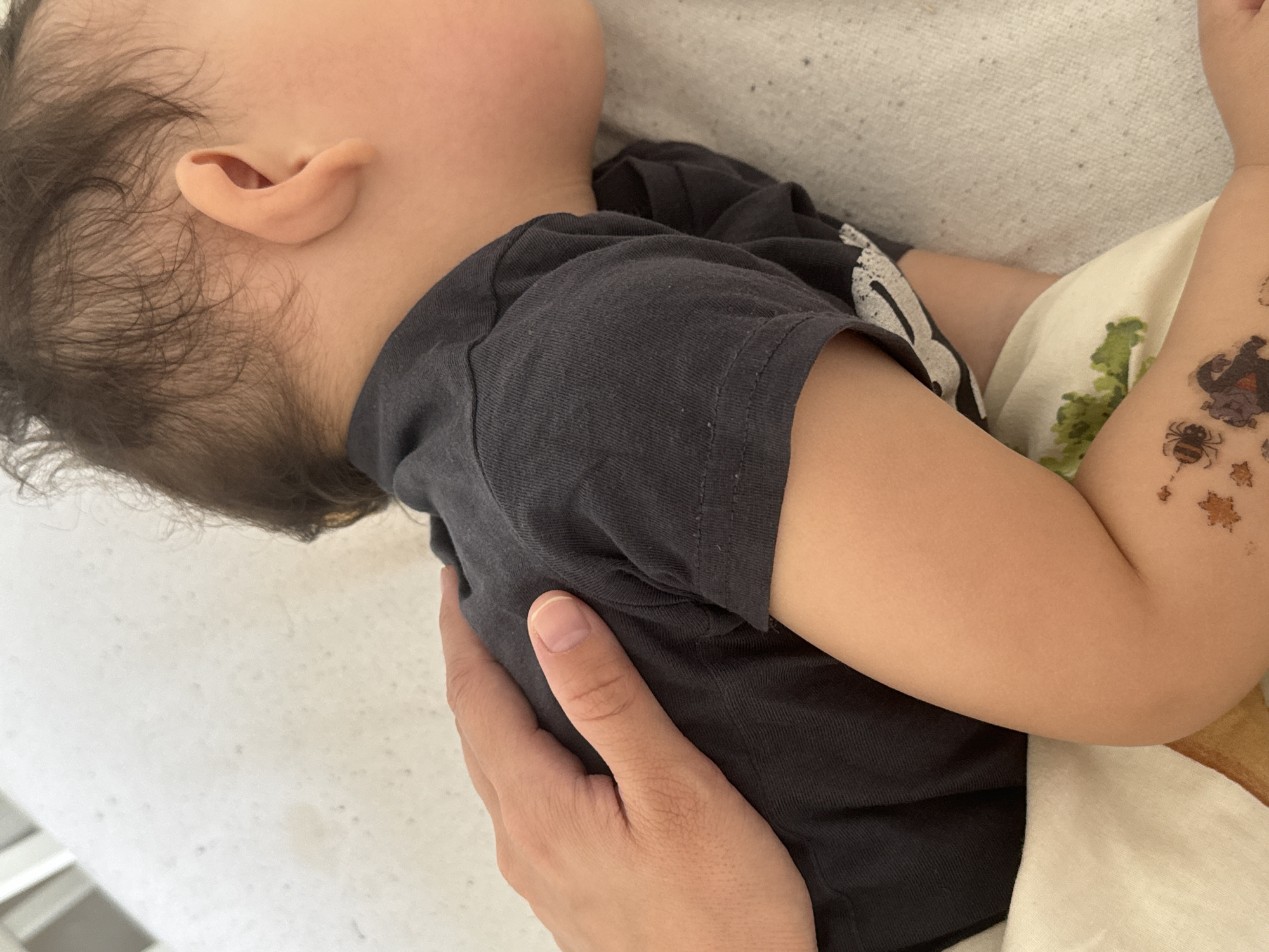 A sleeping child wearing a gray shirt lies on a white textured surface. A human hand rests gently on the child's shirt. The child has light skin and dark hair. There are temporary tattoos on the child's arm. The location is not discernible from the image.