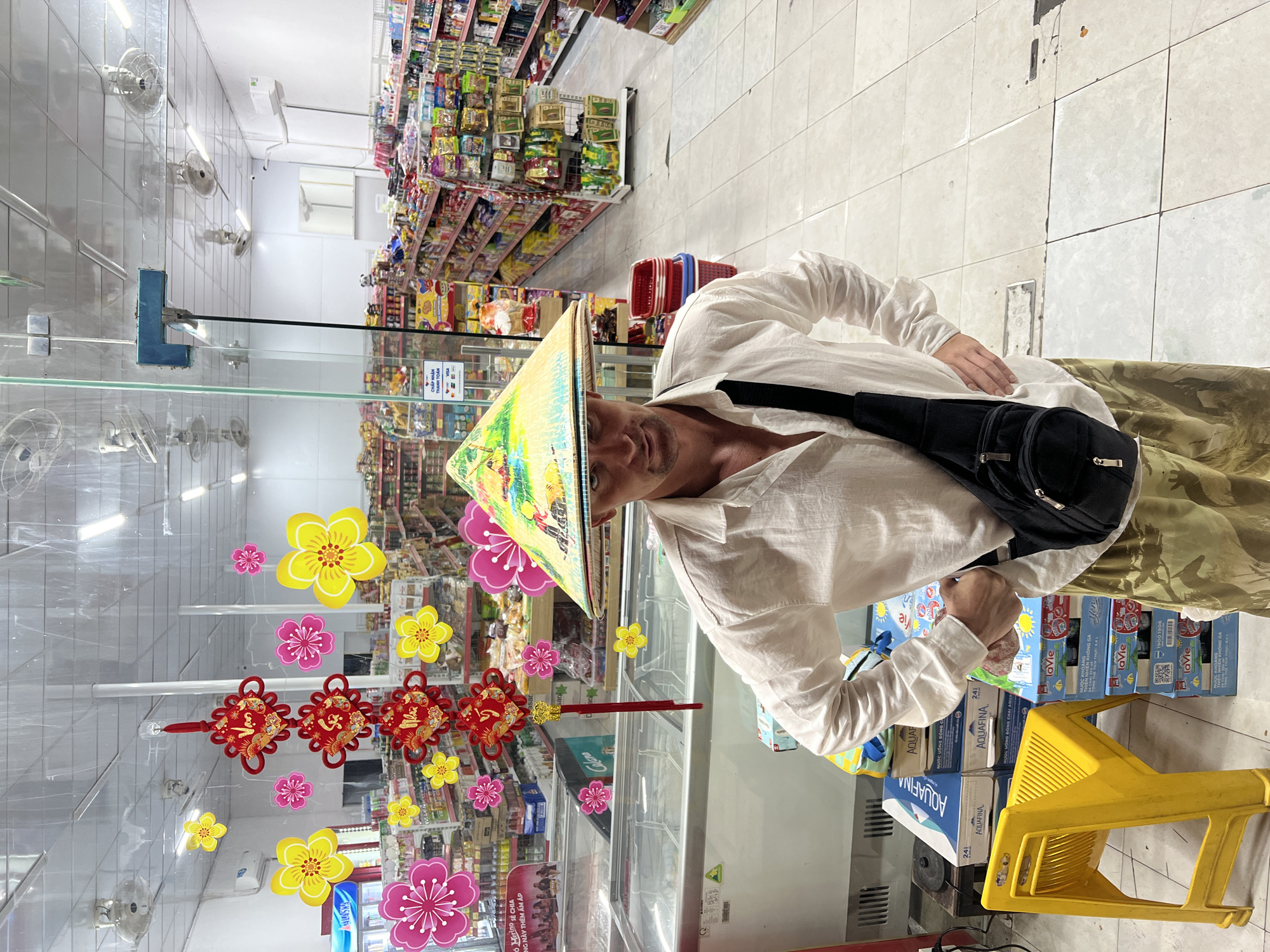 A man wearing a conical hat and a white linen shirt stands in front of a display of goods in a convenience store. The man has a black sling bag across his chest and is wearing camouflage pants. The store is stocked with various products on shelves, and there are refrigerators in the foreground. Red and yellow paper flowers and decorations are hanging in the window of the store, suggesting a festive occasion. The floor is tiled, and the ceiling has fluorescent lighting. The man appears to be looking at the camera. The scene is likely in Đà Nẵng, Vietnam, given the context of the conical hat and store setting.