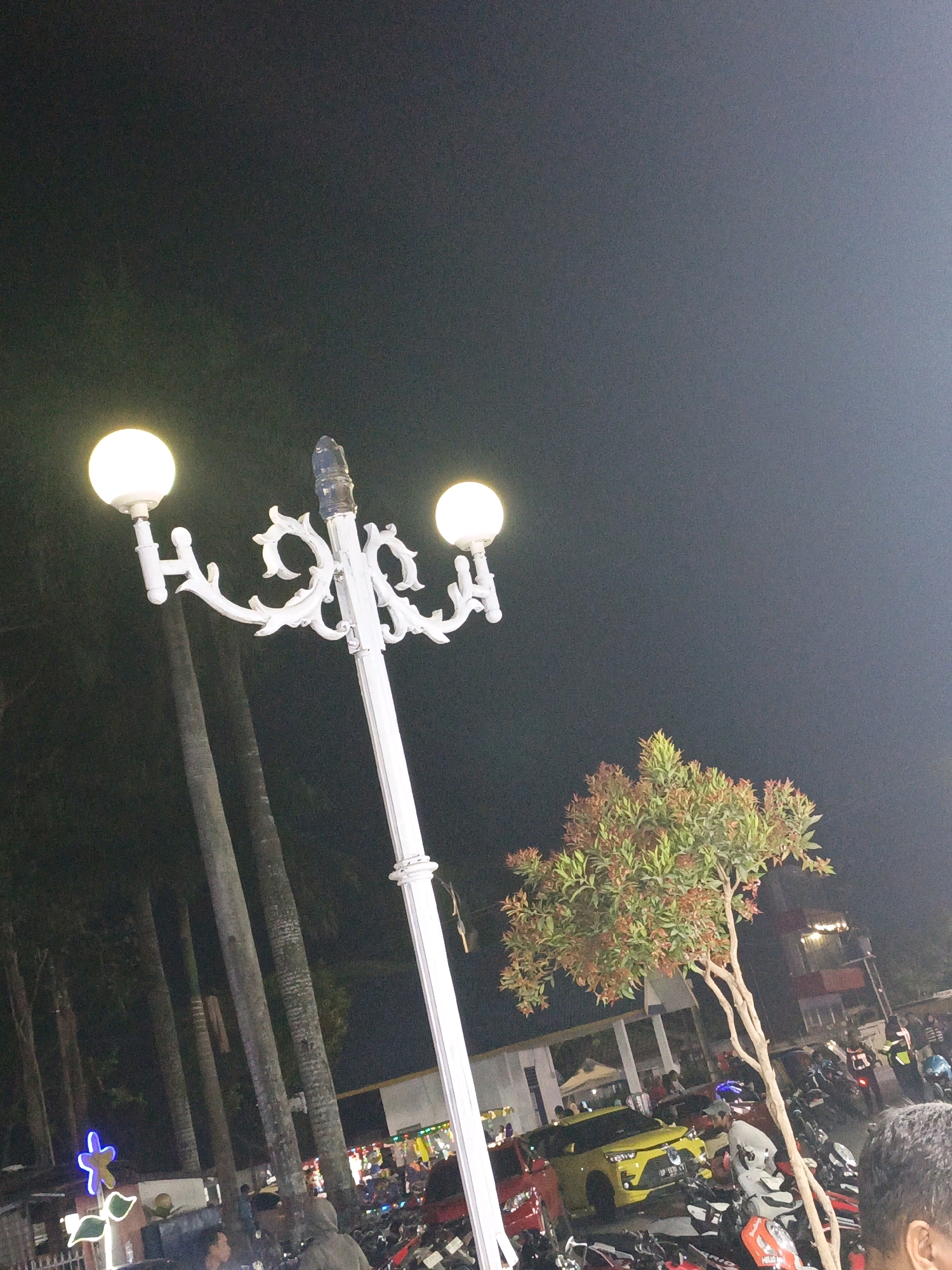 This image depicts a nighttime street scene in Pangkajene, Indonesia. A white ornate streetlamp with two glowing bulbs stands prominently in the foreground, casting light on the surroundings. To the right of the lamp, a small tree with reddish and green foliage is visible. The background is a dark sky. The mid-ground reveals a busy area with parked motorcycles and a yellow car. Several people are present, some standing near the vehicles and others appearing to be walking or gathered. The environment suggests an outdoor gathering or marketplace, illuminated by artificial lighting from the streetlamp and other sources not clearly defined in the image. Tall, slender trees are also visible to the left of the streetlamp, their forms silhouetted against the night sky. In the far background, structures with lights suggest buildings or stalls.