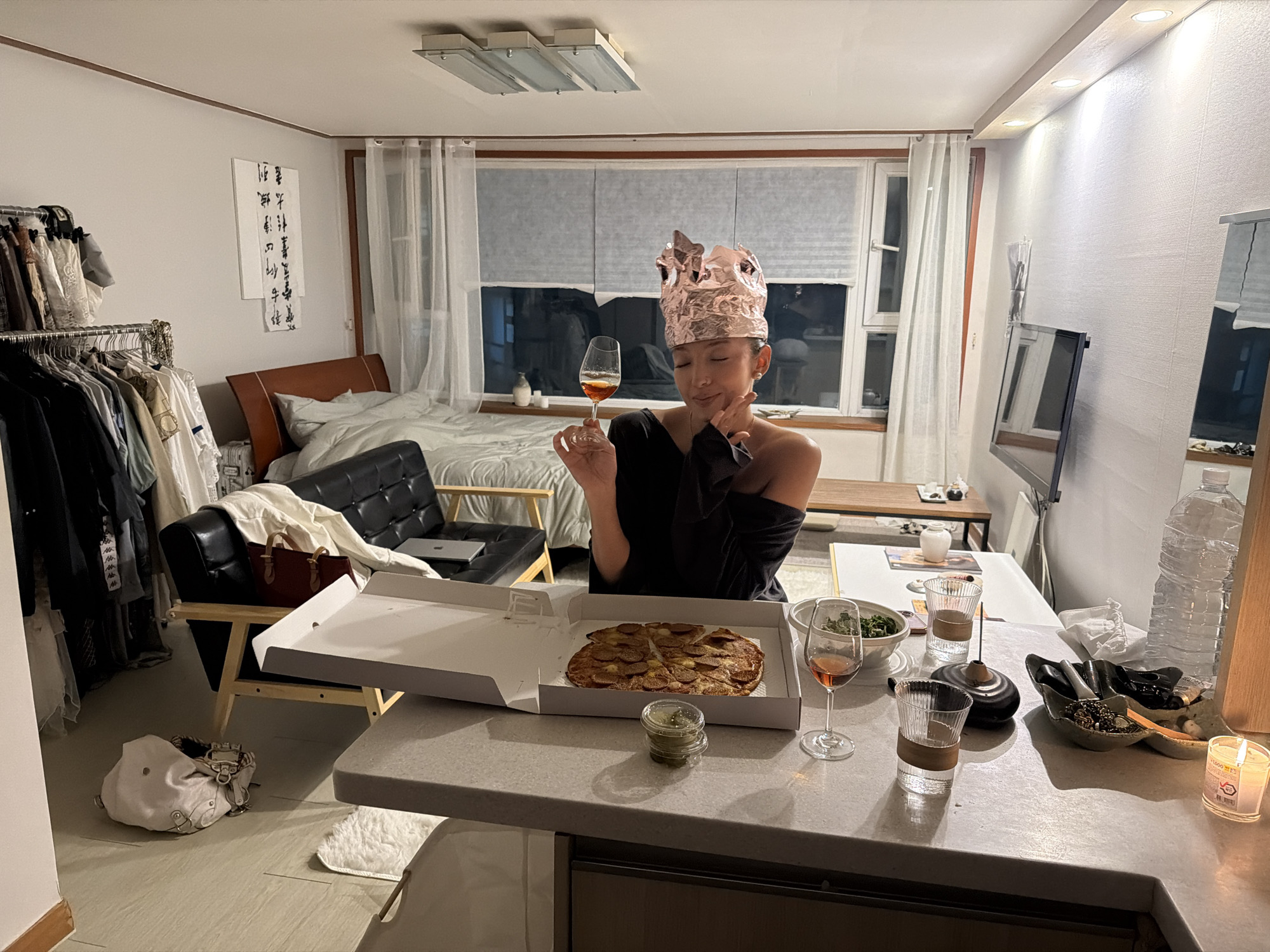 This image captures a cozy and intimate indoor scene at night in what appears to be a compact living space in Seoul, South Korea.
The central subject is a young East Asian woman, her eyes closed in a serene and happy expression, holding a wine glass filled with an amber liquid. She wears a black off-the-shoulder top and a playful, makeshift crown fashioned from crumpled metallic foil.
She is seated at a light grey counter, where an open pizza box with a pepperoni pizza takes prominence. Accompanying the meal are small plastic containers with a green sauce, a bowl of salad, another wine glass, and two clear drinking glasses. The counter also holds a lit candle, an incense holder with a burning stick, a large plastic water bottle, and a small decorative dish containing various small items.
The background reveals a multi-functional room that serves as both a bedroom and living area. To the left, a bed with white bedding and a brown headboard is visible next to a black leather two-seater couch, on which a laptop and a red handbag rest. A clothes rack filled with garments stands near the bed, and a white handbag is on the floor. On the right, a window with white blinds and sheer curtains shows a dark exterior, confirming the nighttime setting. A wall-mounted television or monitor is also present.
The room is illuminated by warm, artificial lighting from overhead fixtures and spotlights. A white vertical artwork adorned with traditional East Asian calligraphy hangs on the wall. The atmosphere feels relaxed and celebratory, possibly marking a casual special occasion or a quiet evening.
Visible text includes illegible East Asian calligraphy on the wall art and the partial number "1600" on the label of the lit candle.