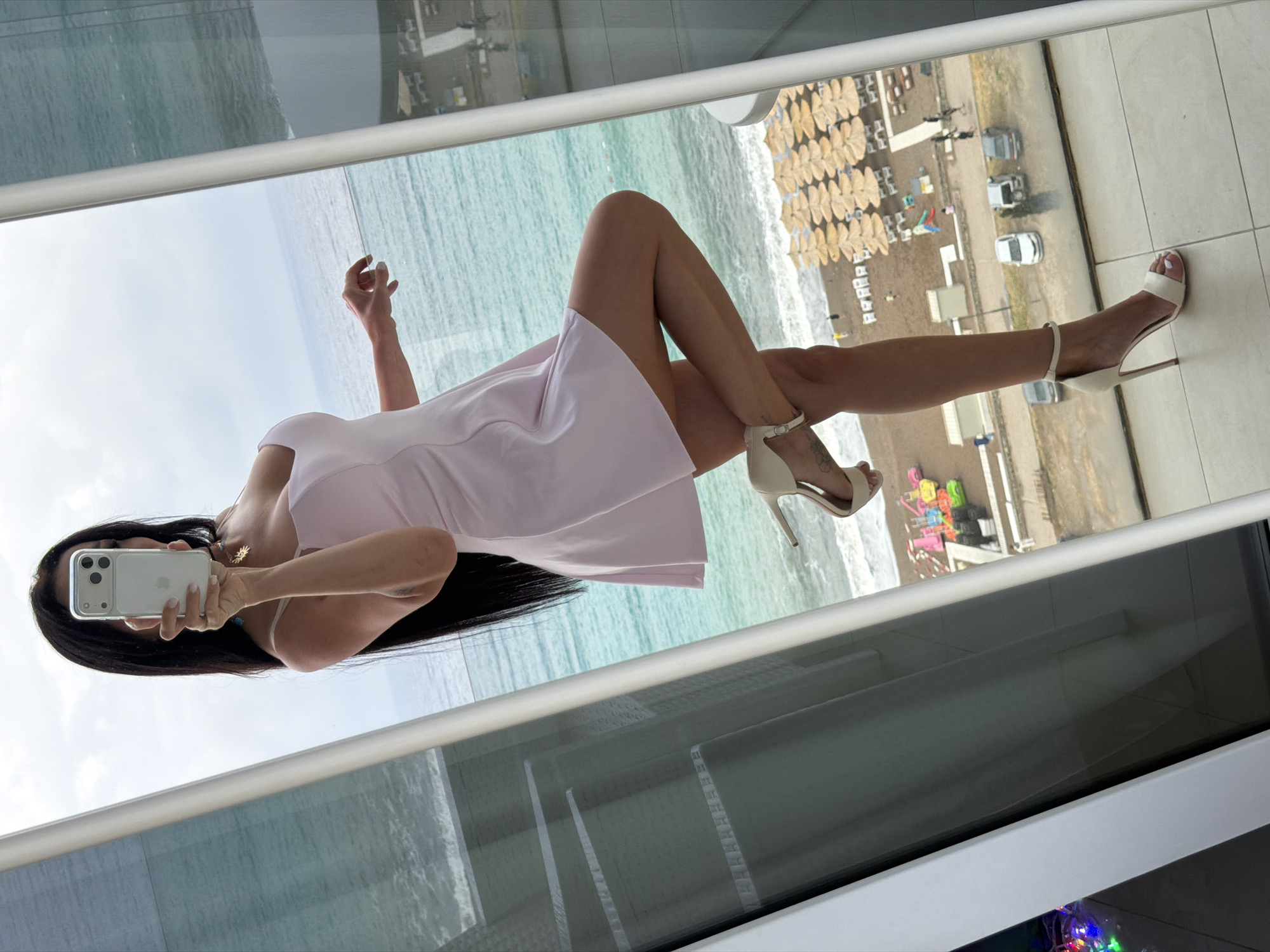 An image rotated 90 degrees counter-clockwise displays a woman lying down and taking a selfie, overlooking a coastal scene from an elevated window. The woman is positioned horizontally across the frame, head to the left, feet to the right. She wears a light pink, possibly sleeveless, dress and white high-heeled sandals with ankle straps. Her long dark hair flows behind her, and her fingernails and toenails are painted white. She holds a white-cased smartphone (likely an iPhone with three cameras) in her left hand, which reflects her face and a necklace with a sun-like pendant.
The background, seen through a large window, reveals an outdoor setting. The upper portion shows an overcast sky, transitioning to the turquoise sea with gentle waves breaking on the shore. Below the sea is a sandy beach, densely populated with rows of straw umbrellas and sun loungers. Several people are visible on the beach, alongside a colorful children's playground structure and a few parked cars on a road running parallel to the coast. Distant buildings line the horizon. The white window frame separates the indoor foreground, where a hint of colorful fairy lights is visible at the bottom right, from the outdoor panorama. The scene depicts a leisure moment at a coastal location in Boreti, Montenegro, during daytime with bright, cloudy weather. No legible text is visible in the image.