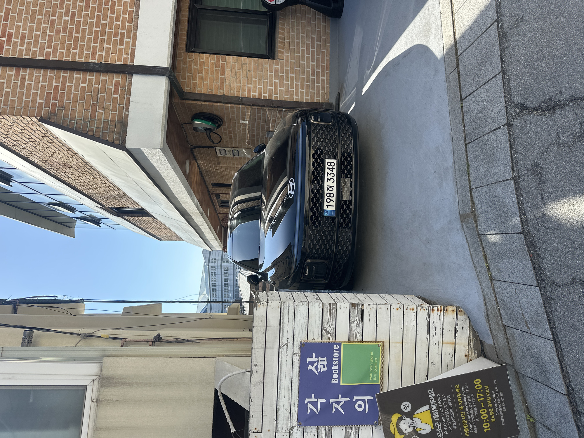 The image shows a black Hyundai car with a license plate reading "198허 3348" parked in a narrow alleyway in Seoul, South Korea. The alley is bordered by brick and plaster buildings. A sign for a bookstore named "각자의 삶" (Gakjaui Salm) is visible, which translates to "Each Person's Life." Another sign requests visitors to keep their voices down and specifies visiting hours from 10:00 to 17:00, noting that the alley is closed on Sundays. The setting appears to be a residential area, likely a hanok village, given the architectural details and the signs emphasizing respect for visiting hours.