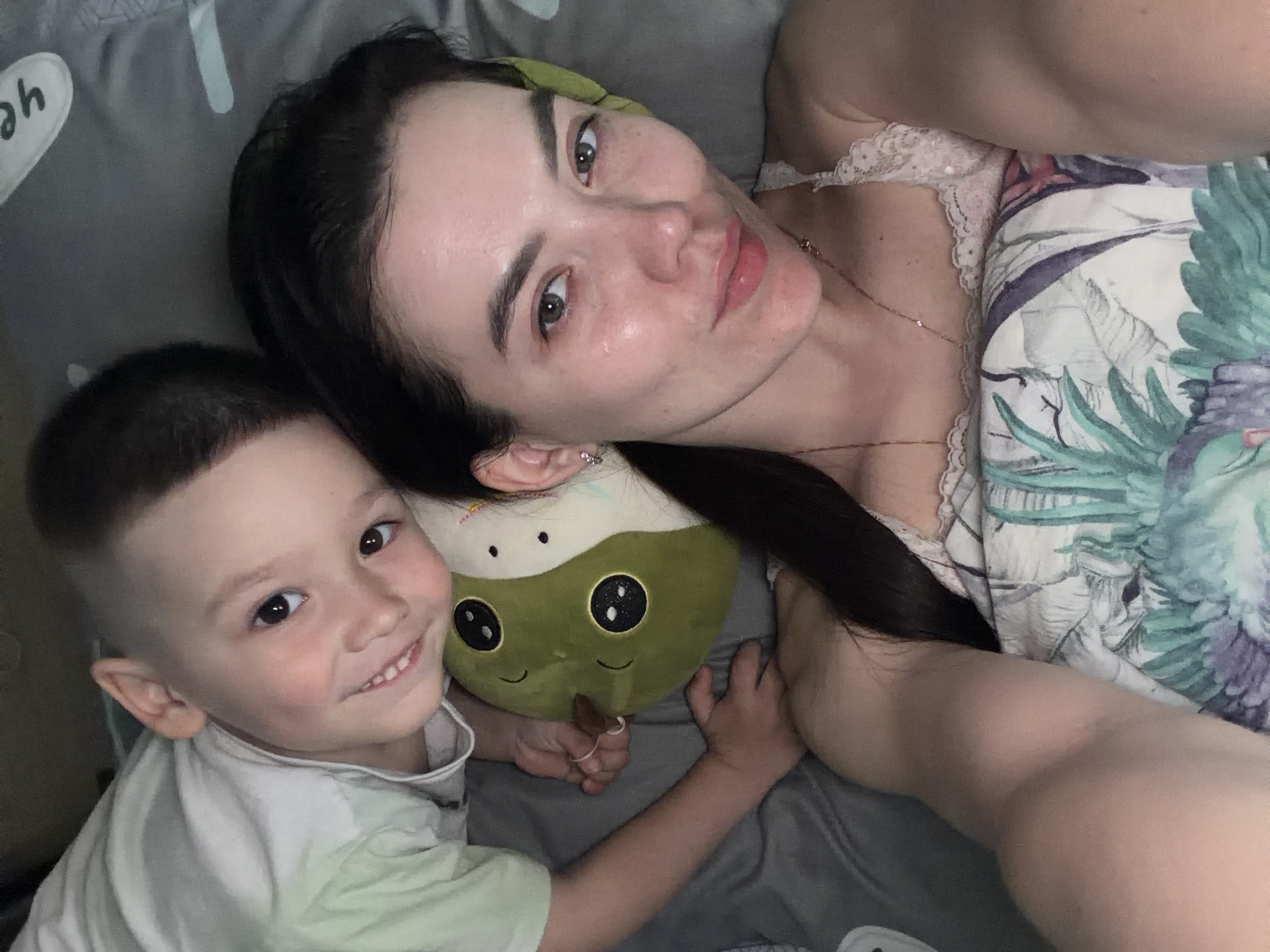 A woman and a young boy are lying on a bed, likely indoors. The woman has long dark hair and fair skin, and she is wearing a light-colored top with lace. The boy has short, light-colored hair and is wearing a light-colored shirt. They are both smiling slightly. Between them is a plush toy. The bed has patterned sheets and a gray blanket. The city location is not discernable from the image.