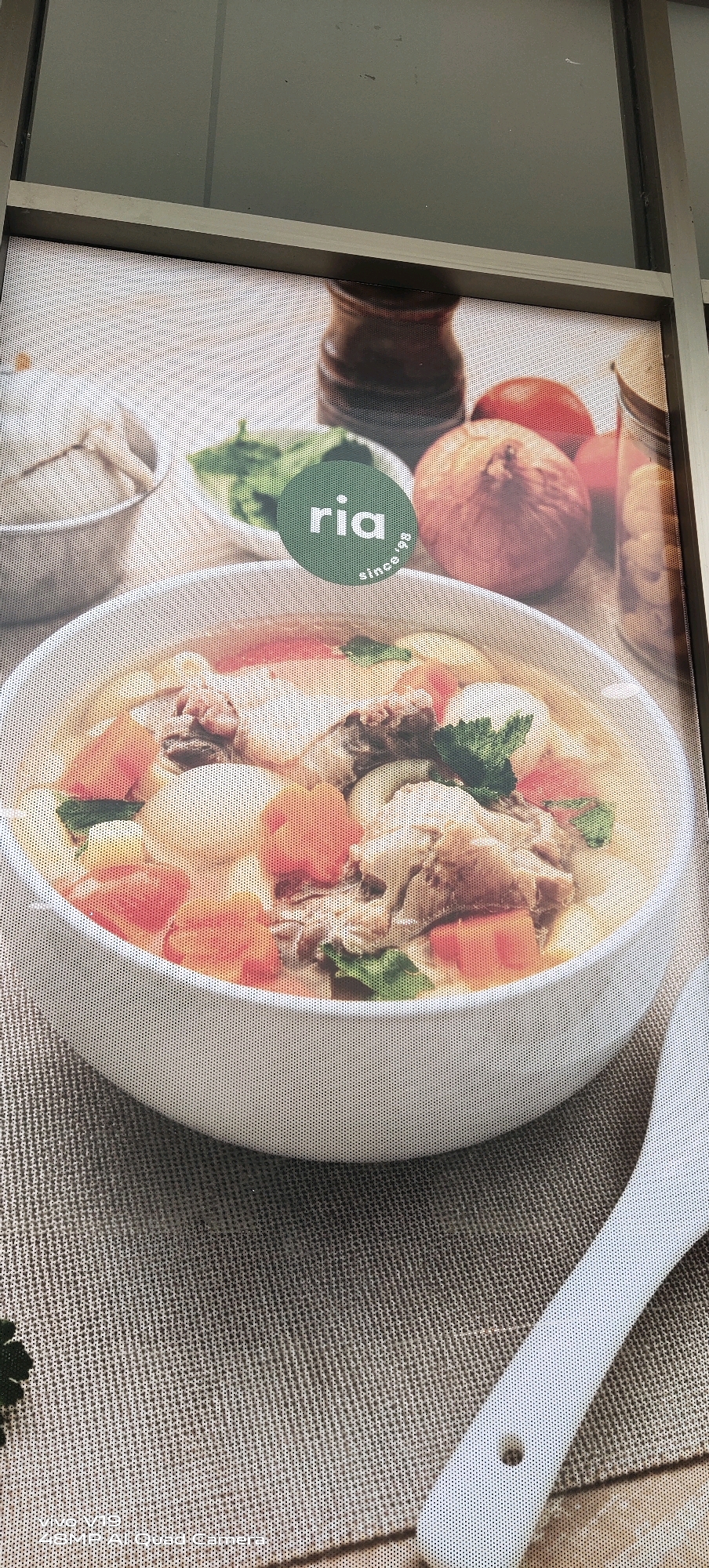 This is a photograph of a storefront advertisement for a restaurant named "ria since 1998". The advertisement features a close-up of a bowl of chicken soup, garnished with herbs. In the background, there are onions, tomatoes, and a grinder. The advertisement is mounted on a transparent surface, possibly a window, with the faint reflection of an outdoor environment visible. A portion of a white ladle is also visible in the lower right corner. The advertisement is likely located on a commercial street in Pekanbaru, Indonesia.